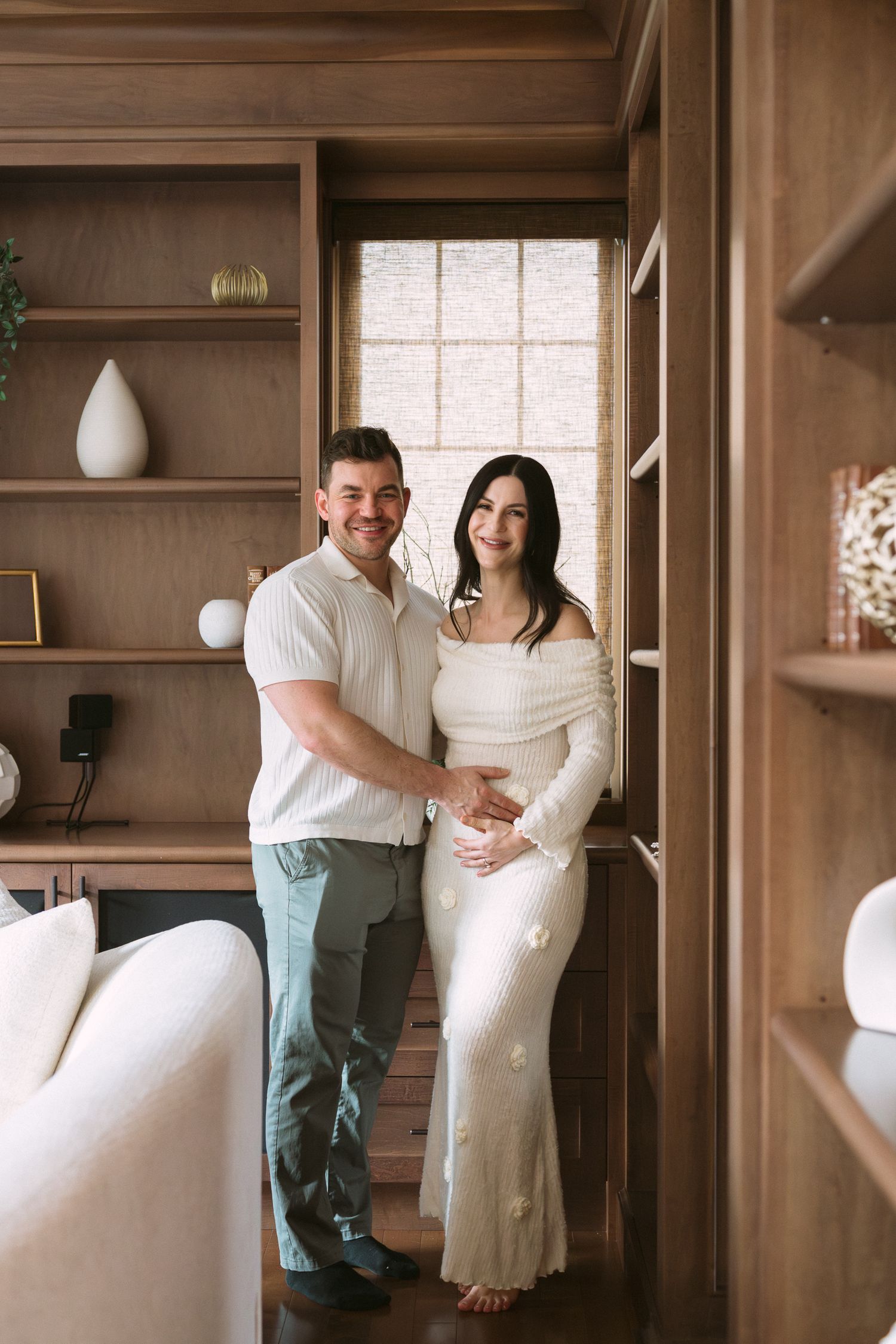 Smiling couple in white outfits pose in a wood-paneled library, man embracing pregnant woman in off-shoulder knit dress.