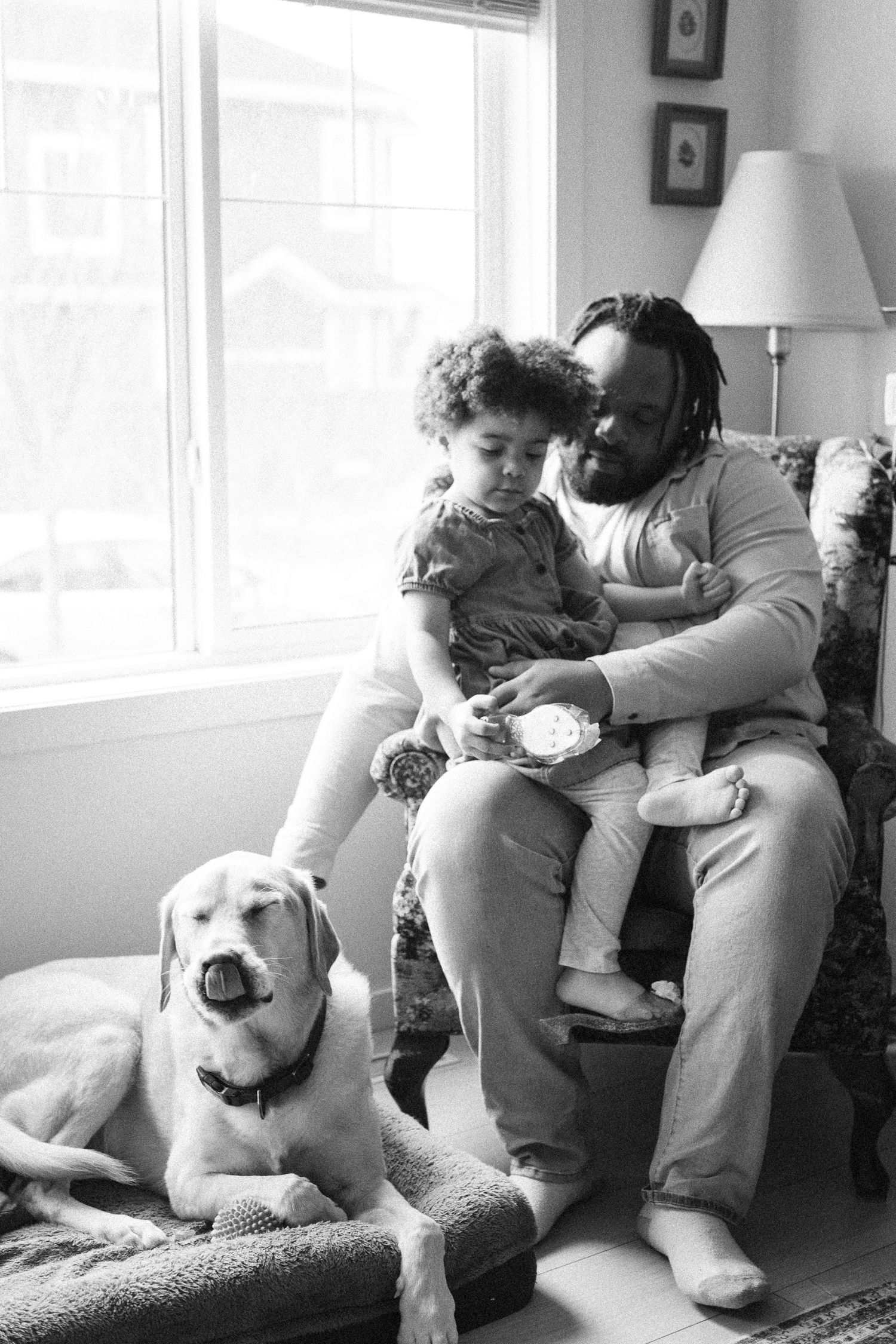 Black and white photo of a bearded father with dreadlocks holding his young child in a chair, with a Labrador dog nearby.