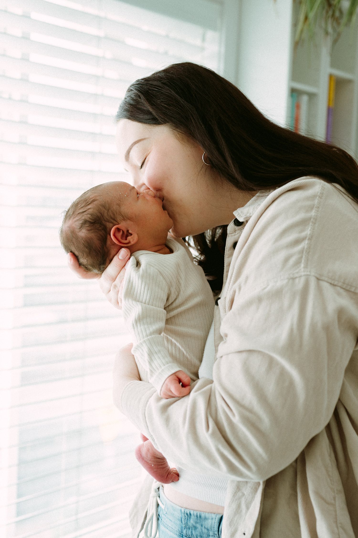 A mother tenderly kisses her newborn baby while holding them gently near a bright window.