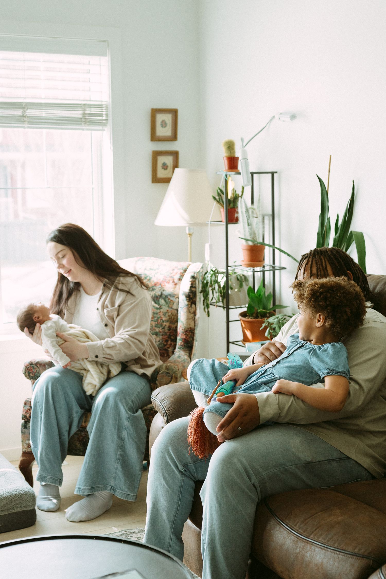 Two women sit on a couch in a cozy living room, one holding a newborn baby and the other cradling a young child.