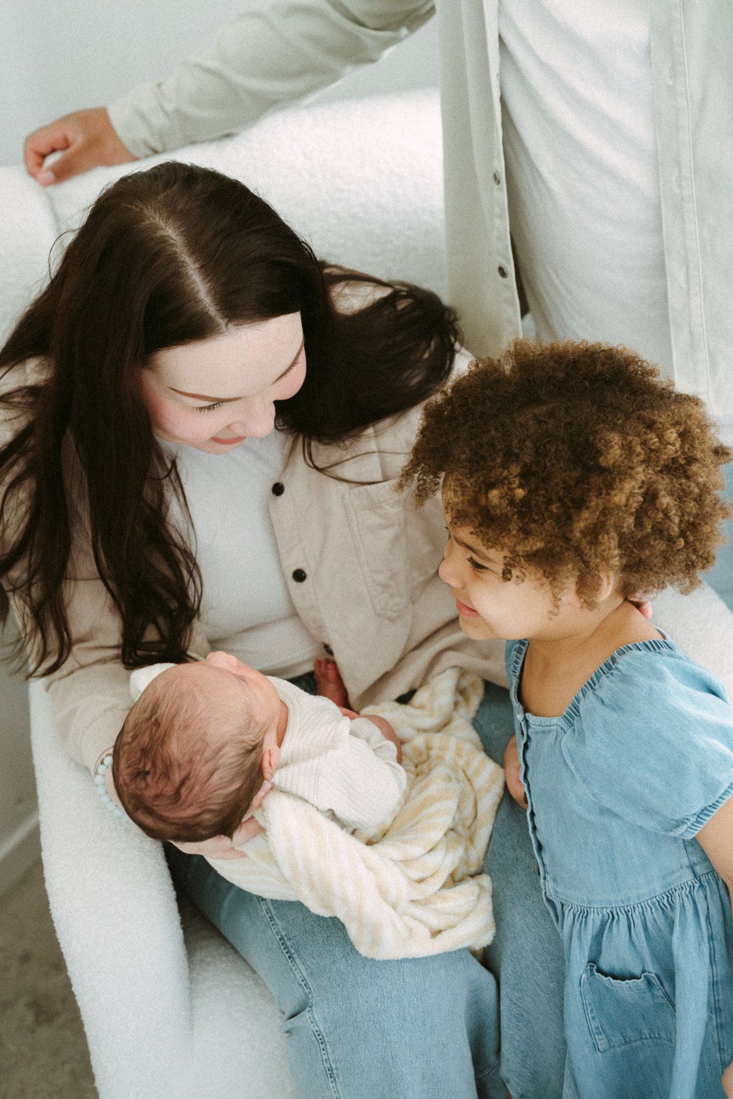 A woman cradles a sleeping newborn in a white chair as a young curly-haired girl in a denim dress gazes lovingly at the baby.