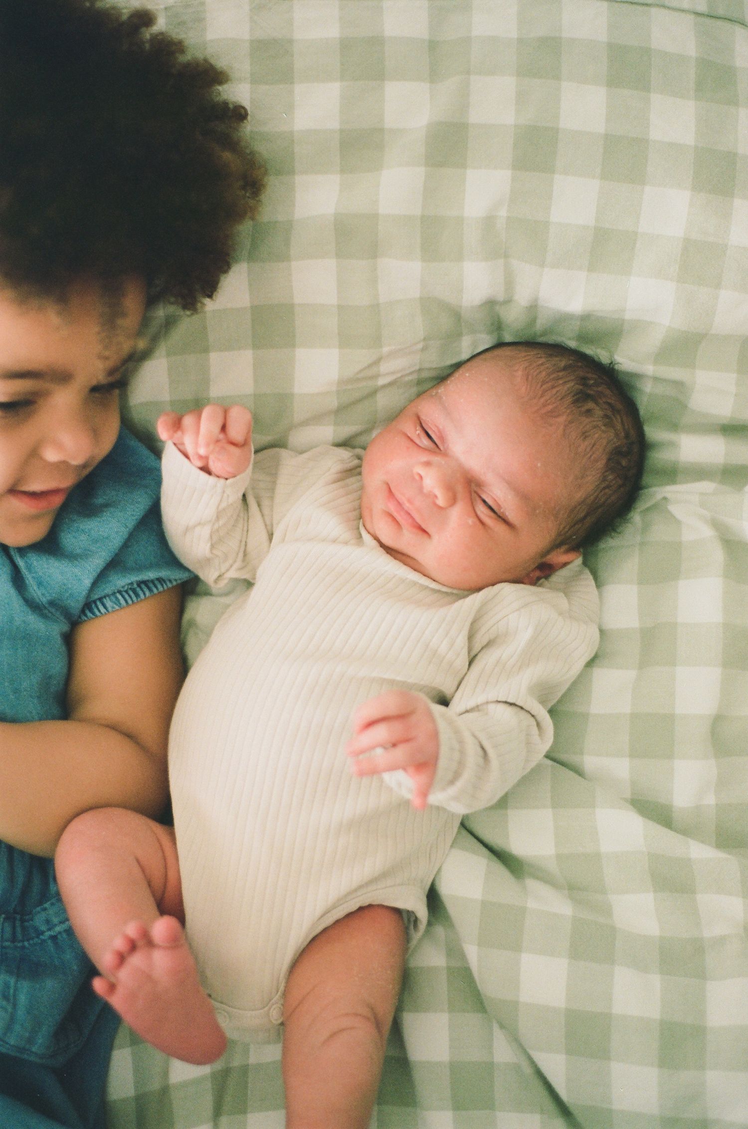 A curly-haired toddler lies beside a smiling newborn baby on green gingham bedding.