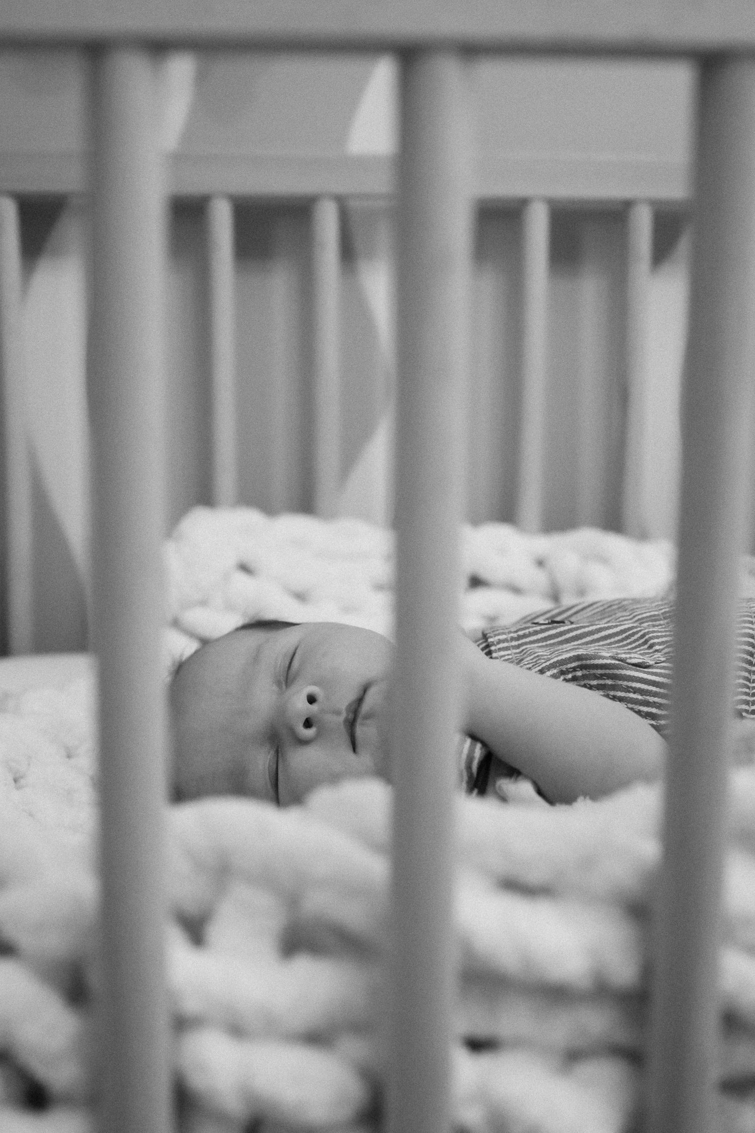View through crib bars of sleeping baby on white bedding in soft lighting.