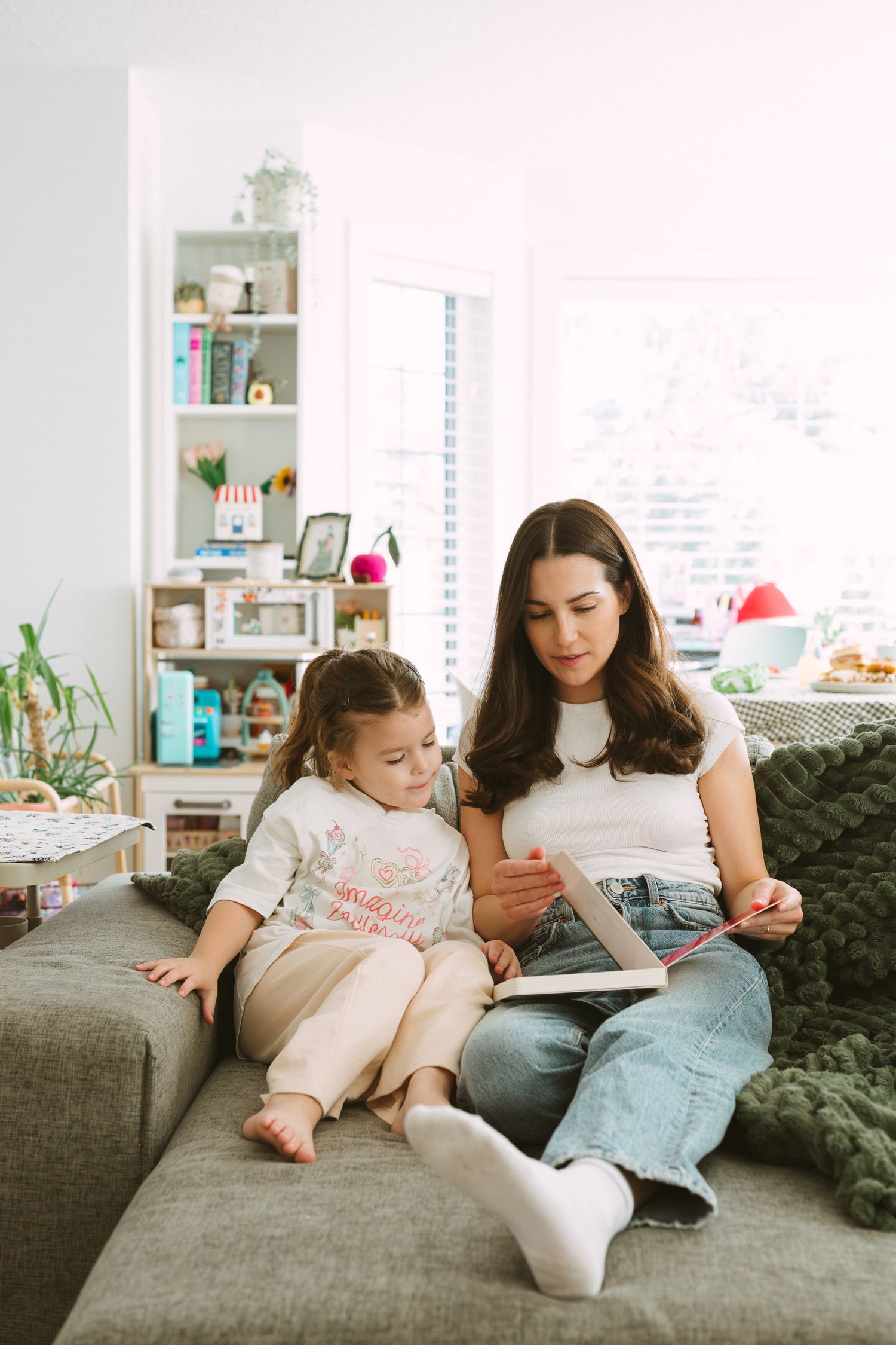 Two people lounge casually on a gray sofa while reading together in a sunlit room with bookshelves.