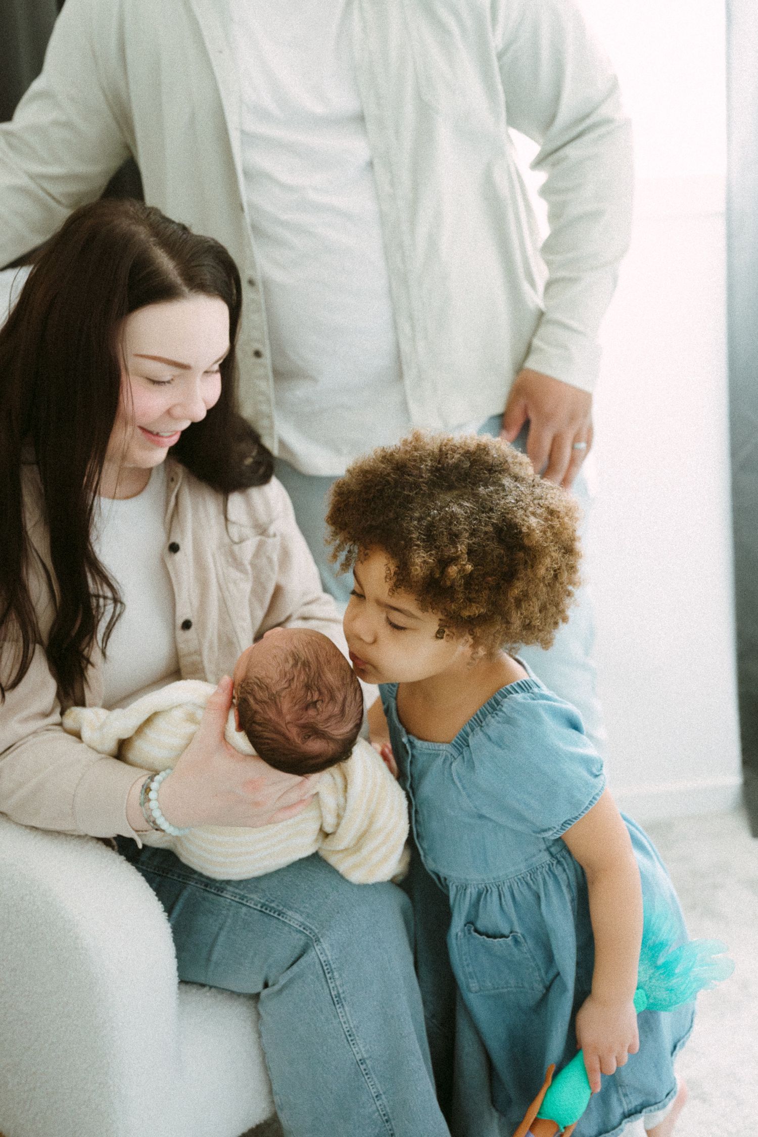 A toddler girl in a blue dress kisses a newborn baby held by her smiling mother.