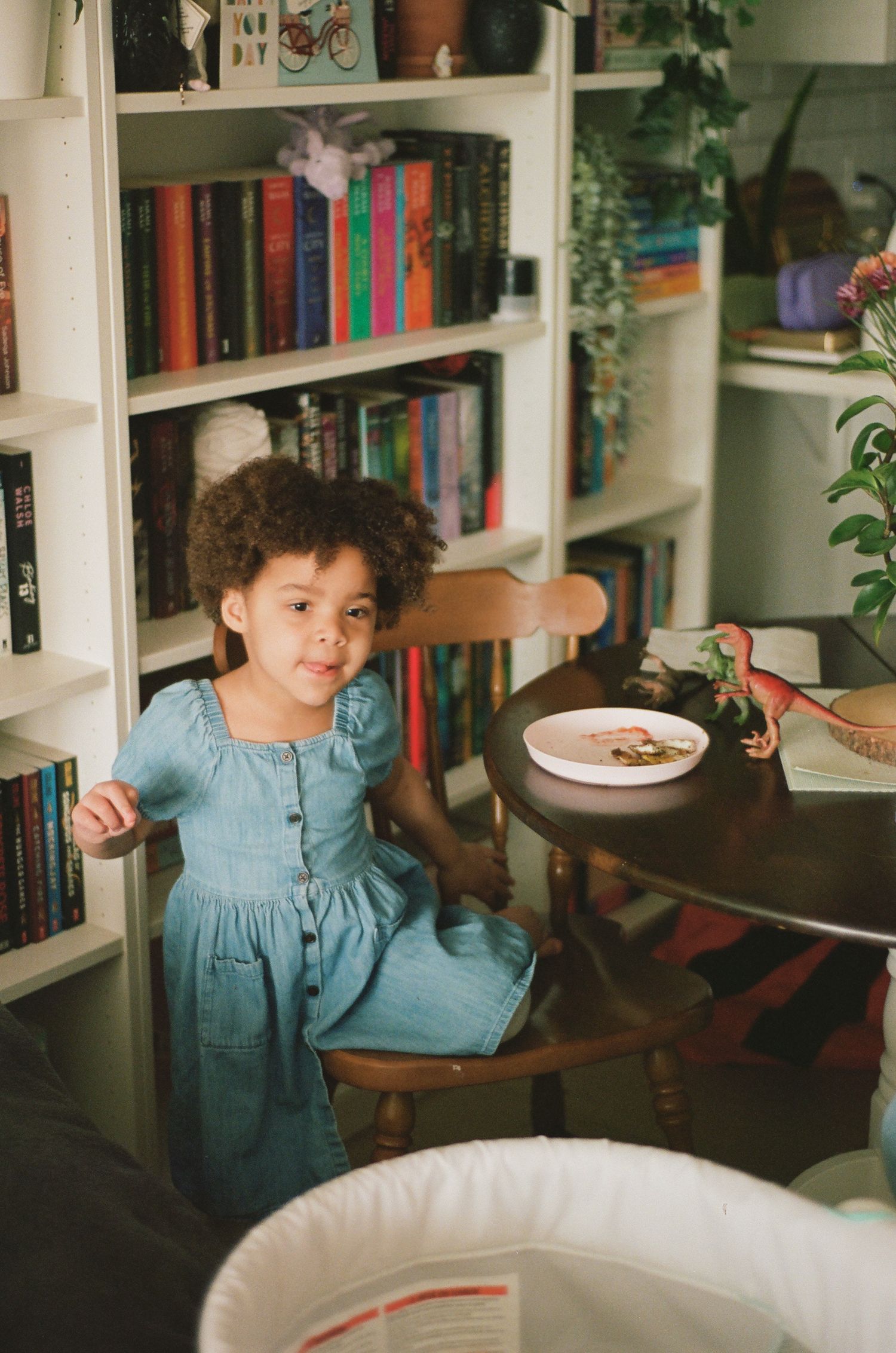 Young girl in blue dress standing by a round table with bookshelf background in cozy home library.