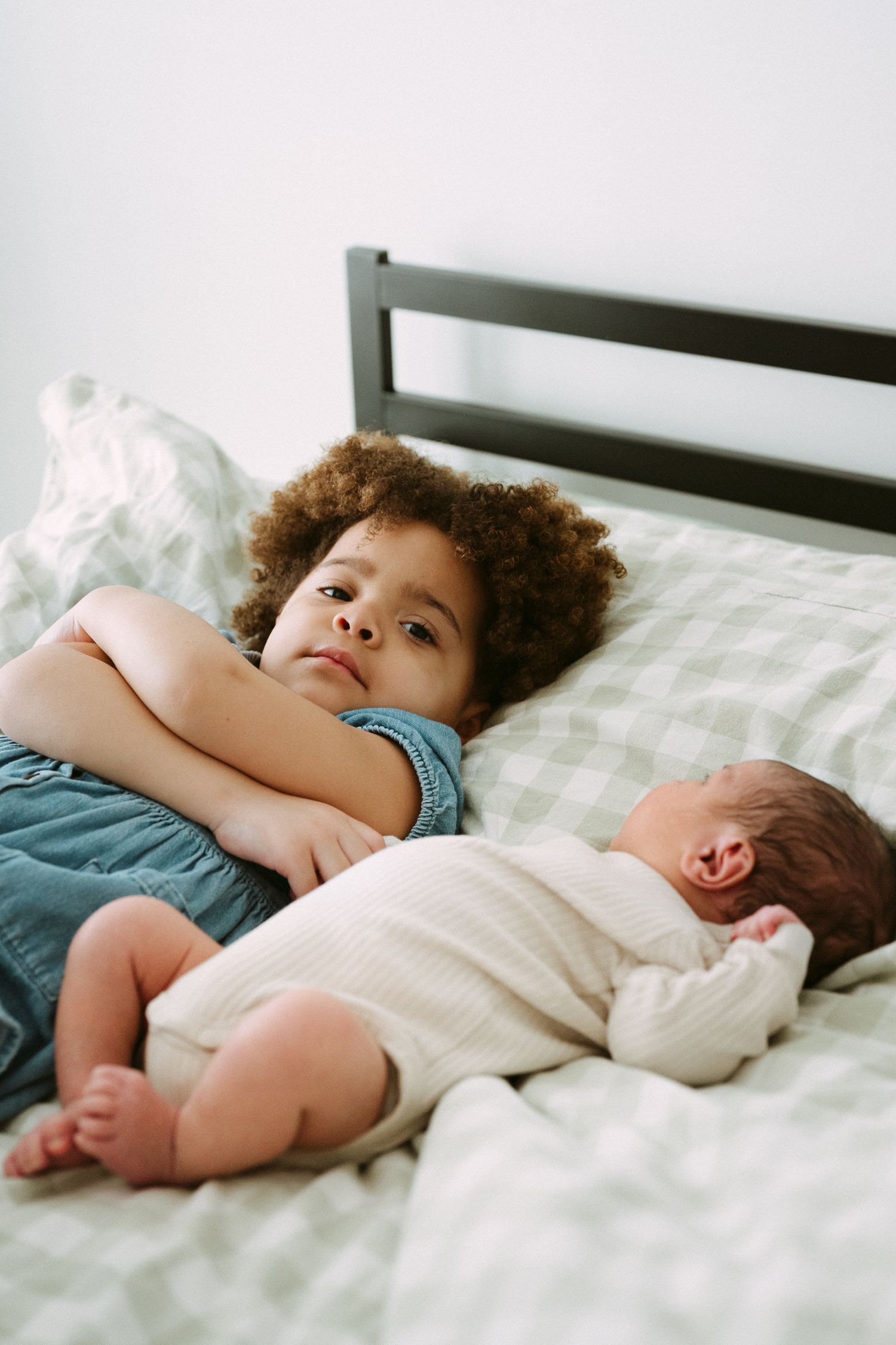 Young child with curly hair lying on bed next to sleeping newborn baby sibling on checkered bedding.