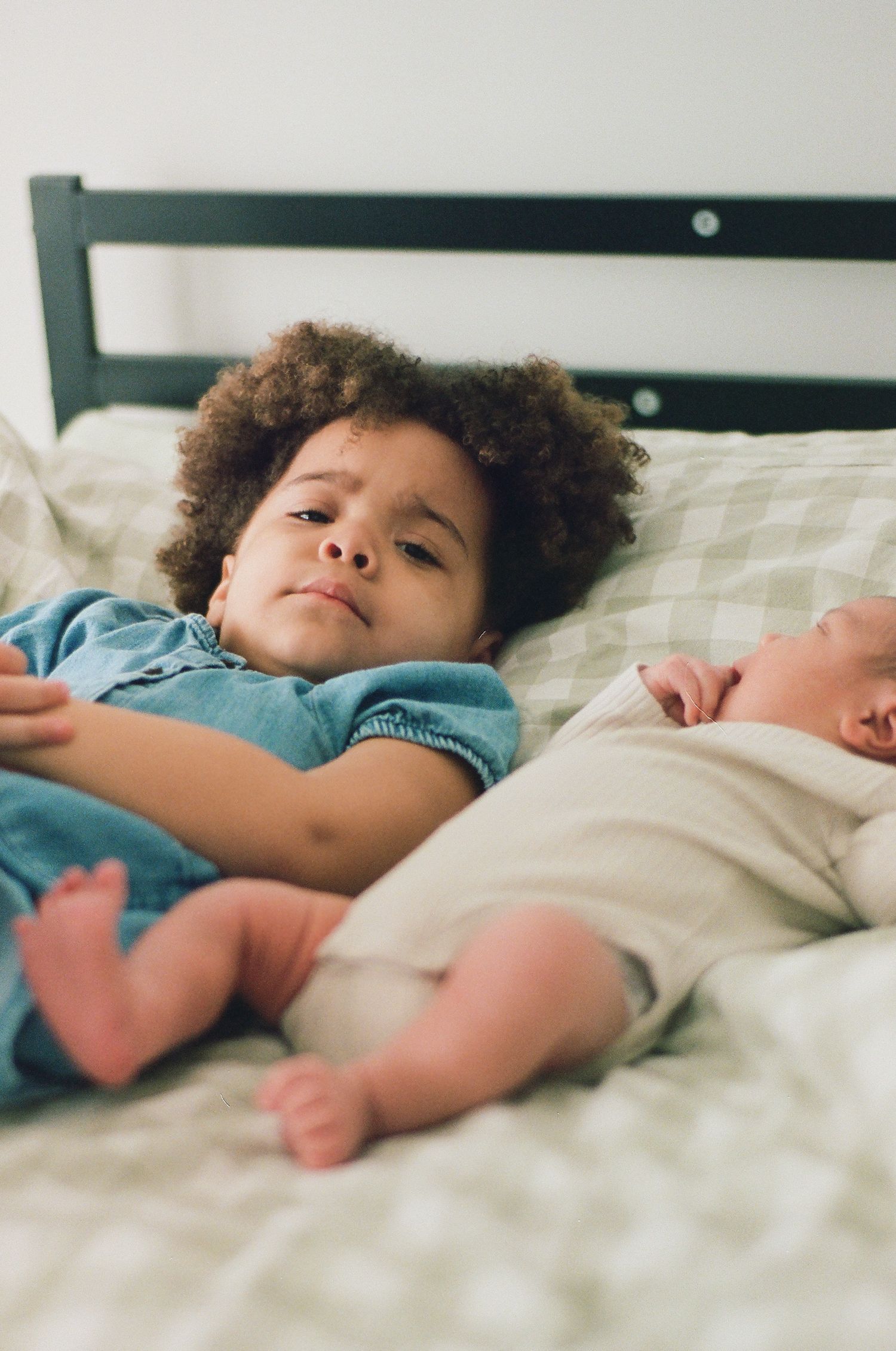 A toddler with curly hair in a blue outfit lies on a bed next to a sleeping newborn baby sibling.
