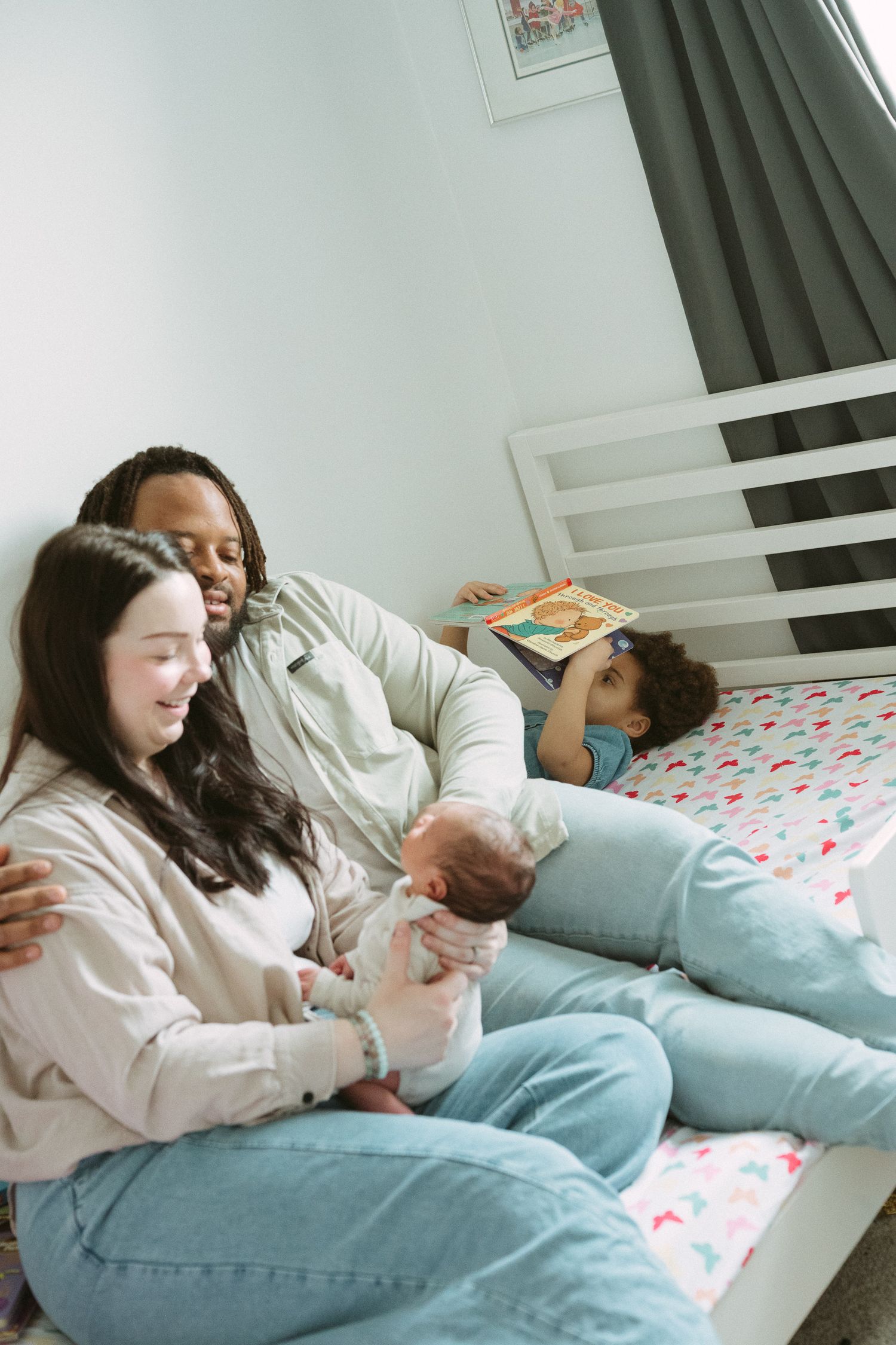 A family sits together on a bedroom floor, parents holding a newborn baby while a child reads a book on the bed.