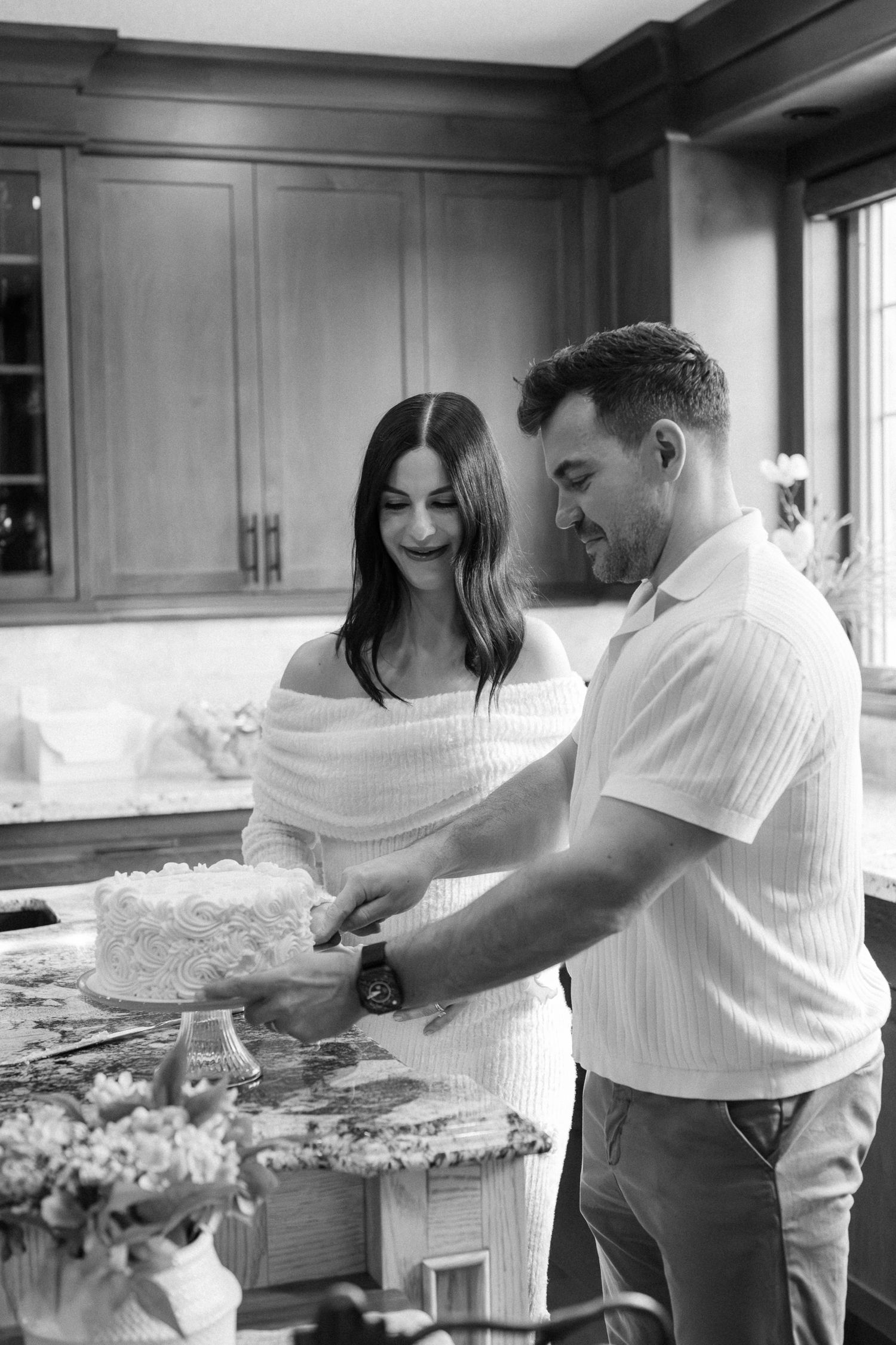 Black and white photo of a couple cutting a white layered cake together in an elegant kitchen.