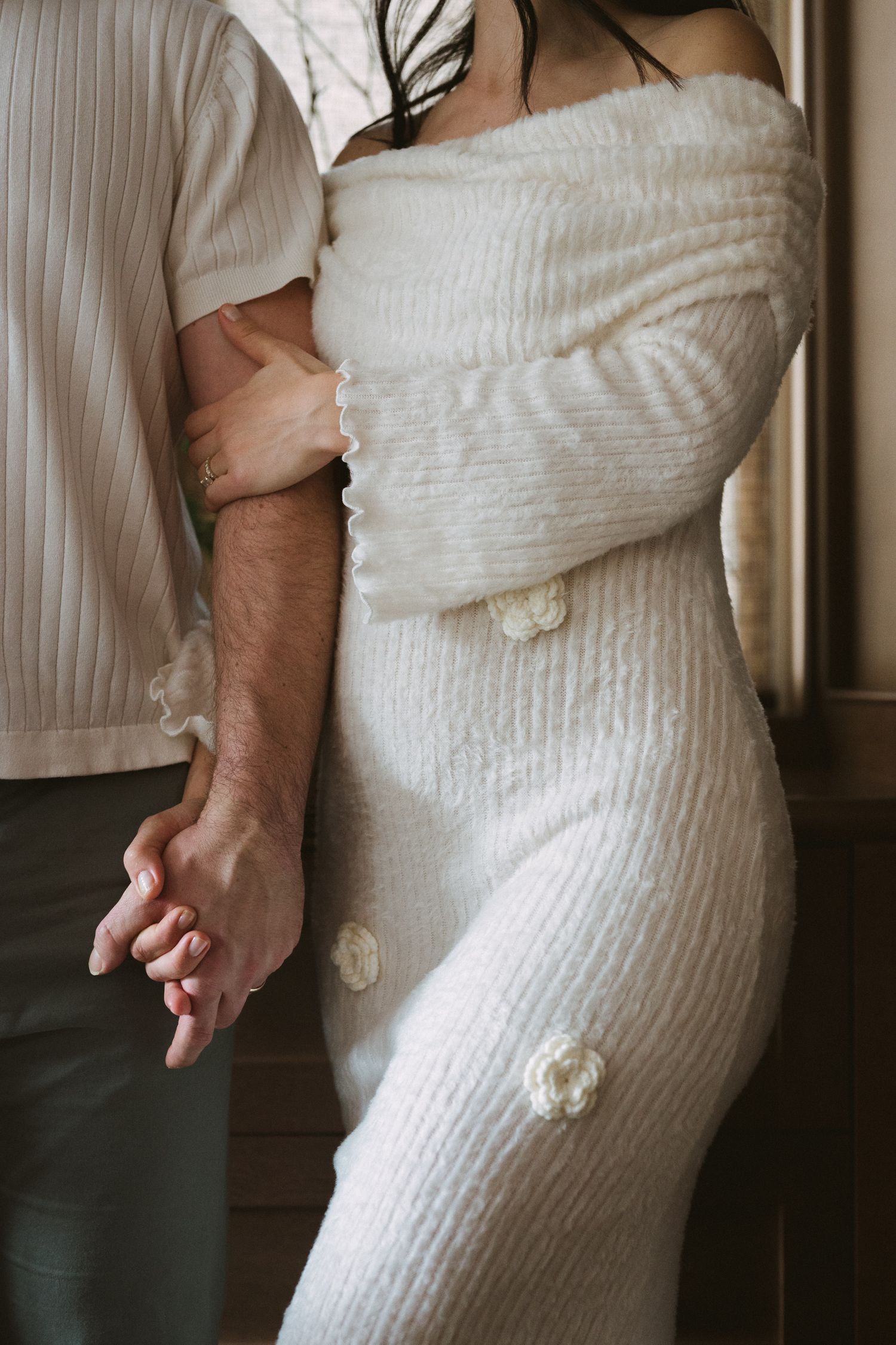 Couple holding hands, pregnant woman in white off-shoulder knit dress with floral details embraced by partner.