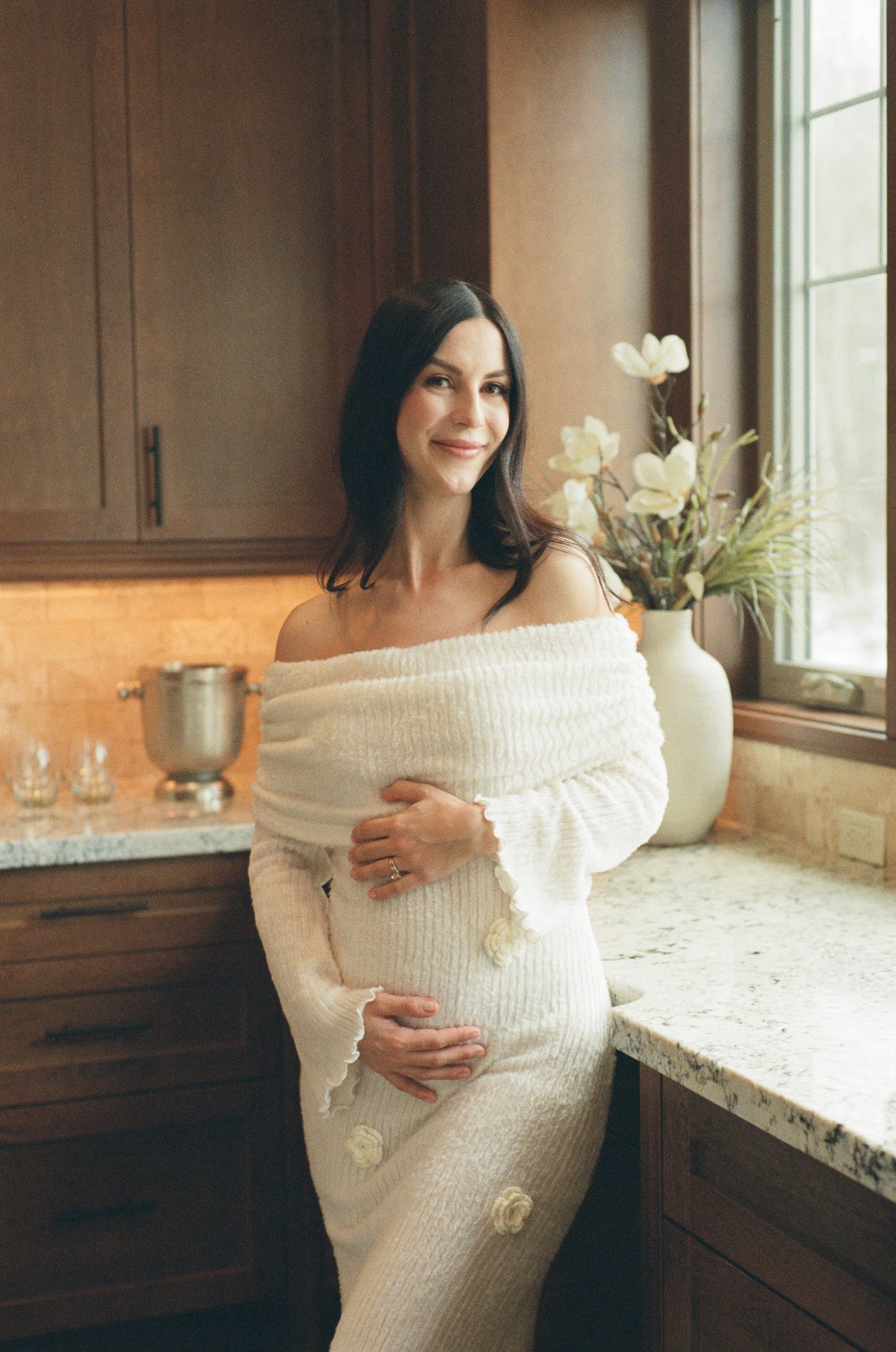 Glowing pregnant woman in white off-shoulder knit dress smiling in kitchen beside vase of white flowers.