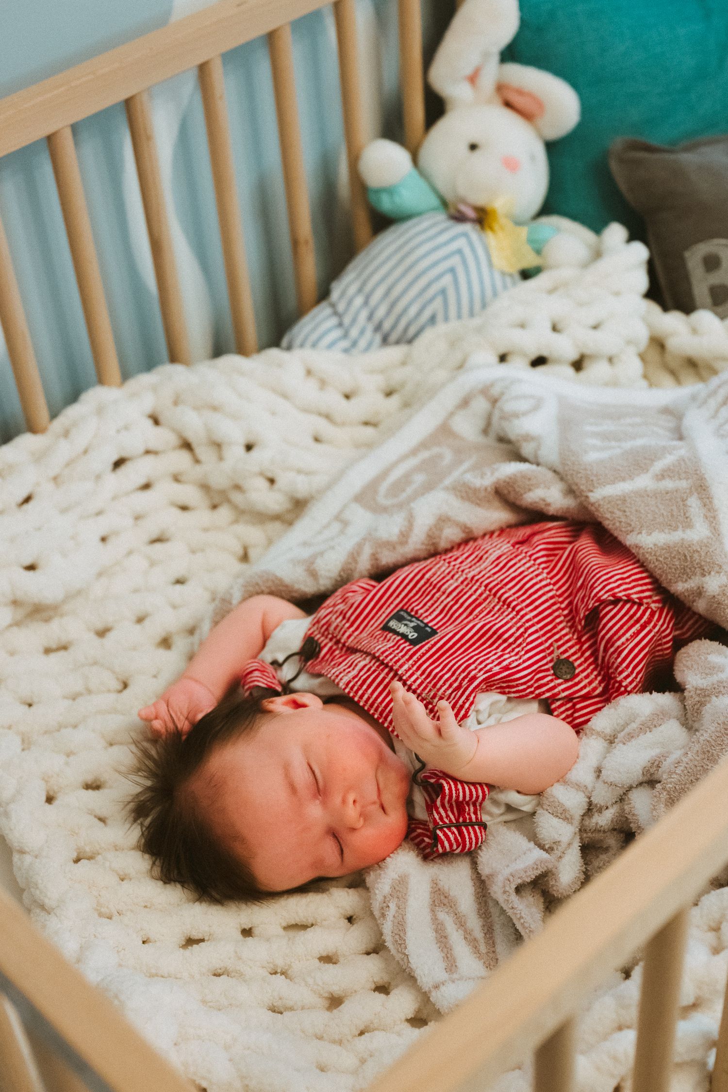 A baby naps in a wooden crib with a chunky knit blanket and stuffed animals nearby.