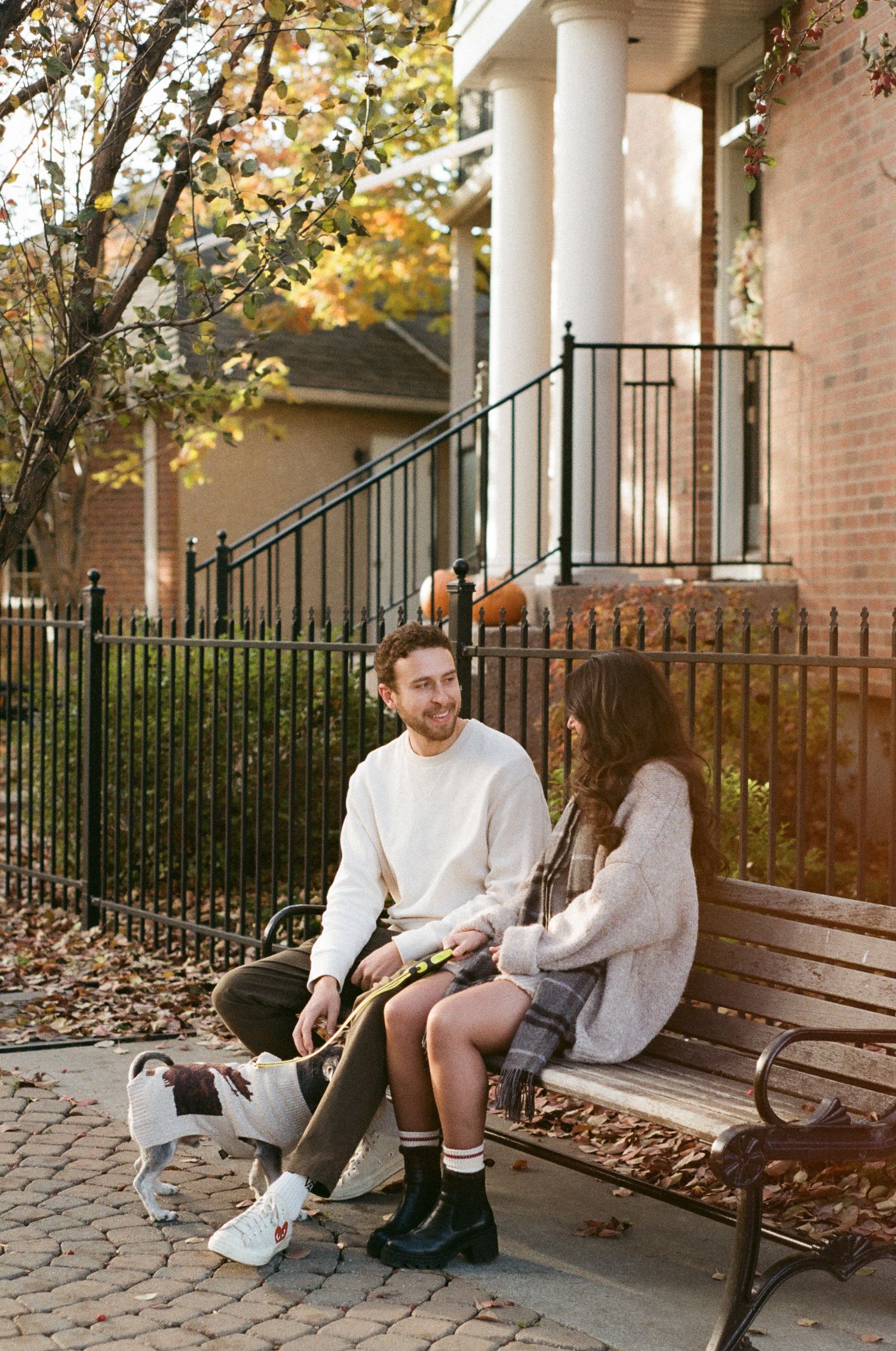 A couple sits together on a bench under white columns with fall foliage in the background.