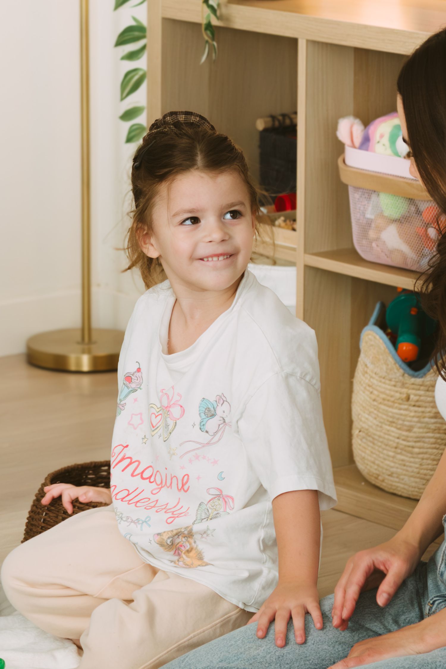 A child wearing a white t-shirt sits and smiles in a playroom with storage shelves and baskets in the background.