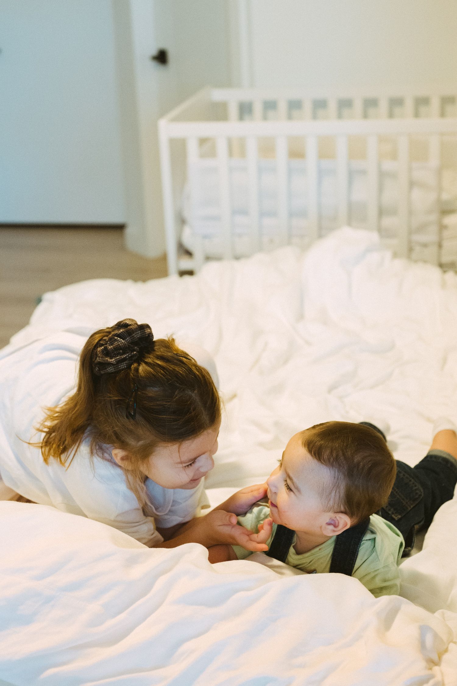 Two children share a sweet moment while lying on white bedding in a bright room.