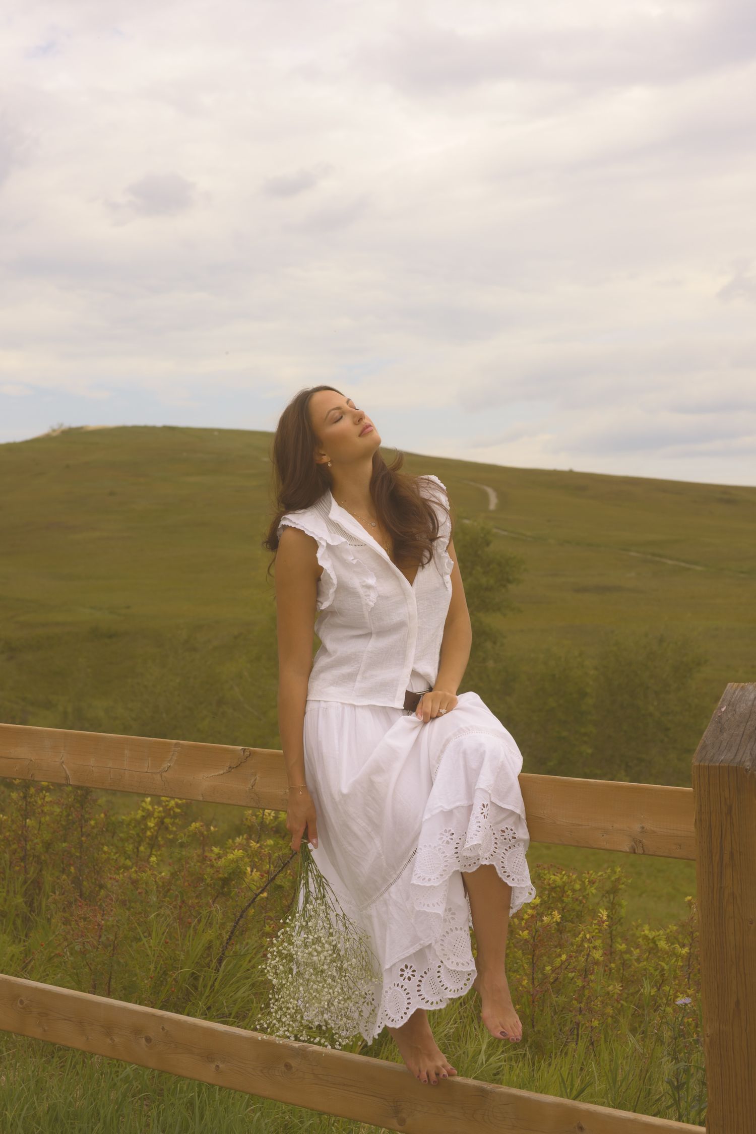 A whimsical scene captures movement in a white dress while sitting on a rustic fence.