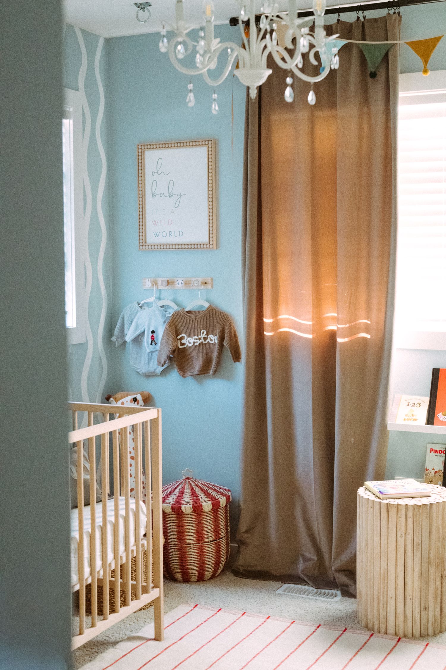 Elegant nursery with chandelier, natural wood crib, and blue walls with brown curtains.