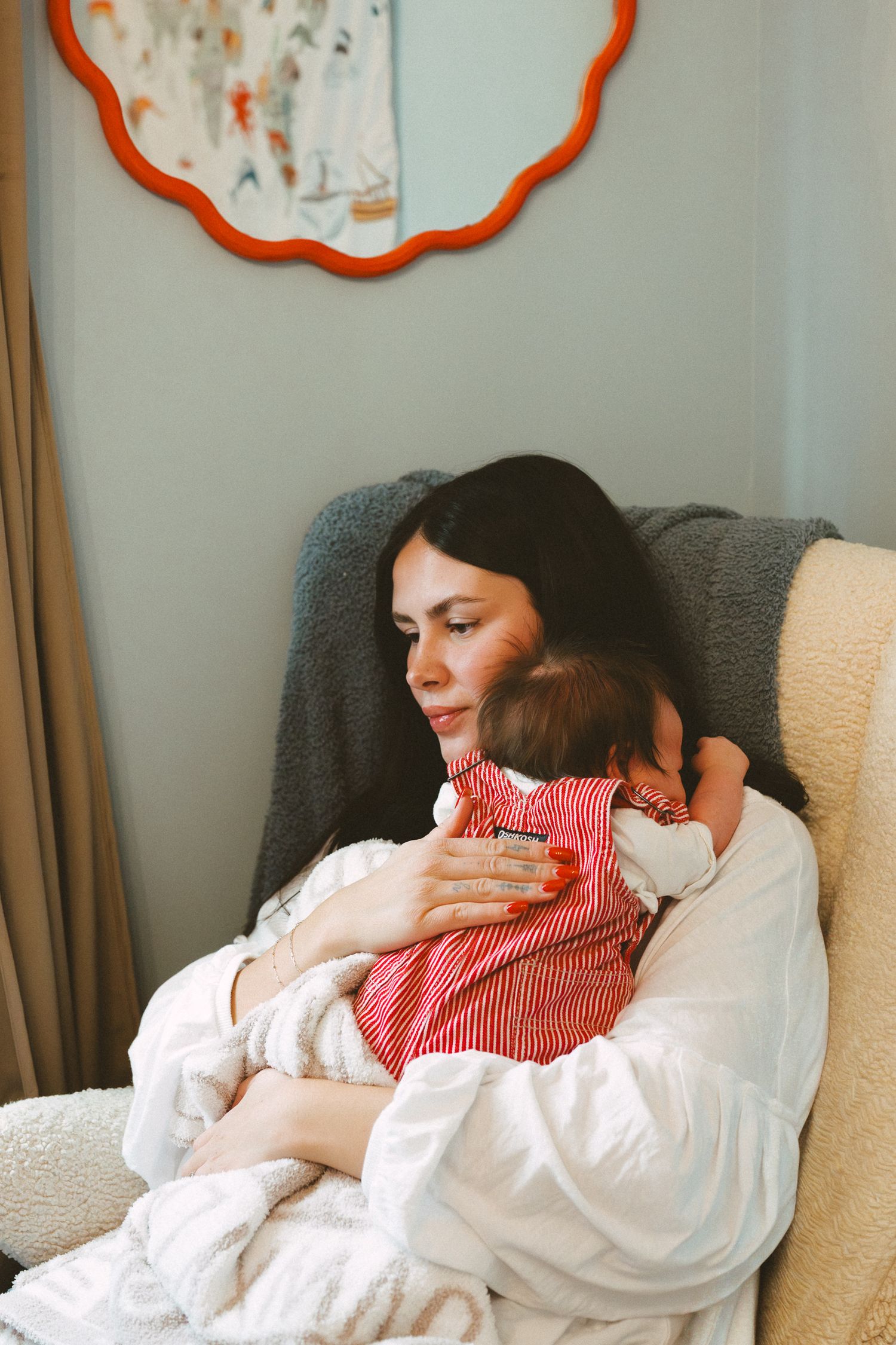 A parent in a white blouse holds a sleeping baby wearing red stripes while resting in a cozy gray armchair.