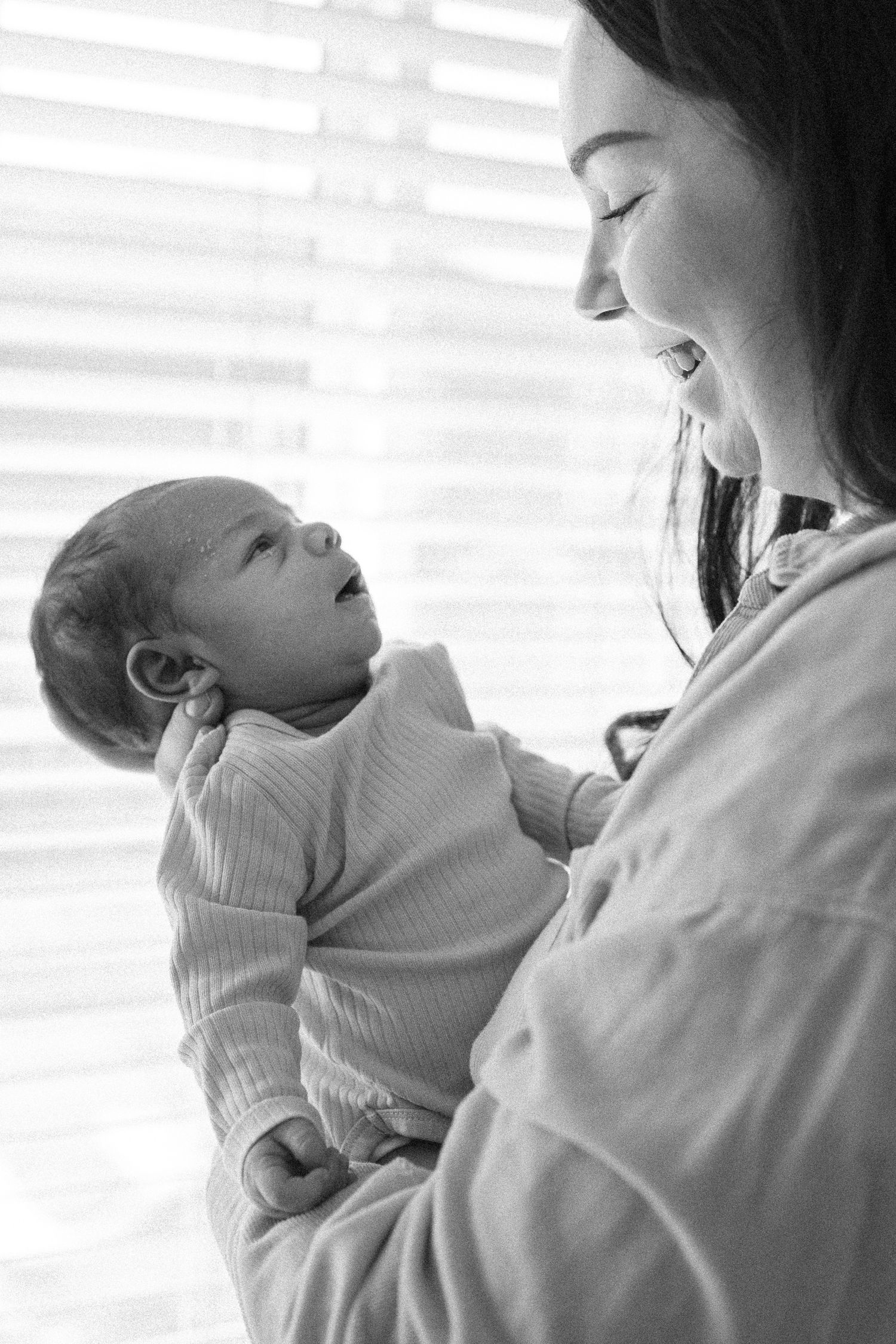 Black and white photo of a smiling mother tenderly holding her newborn baby near a bright window.
