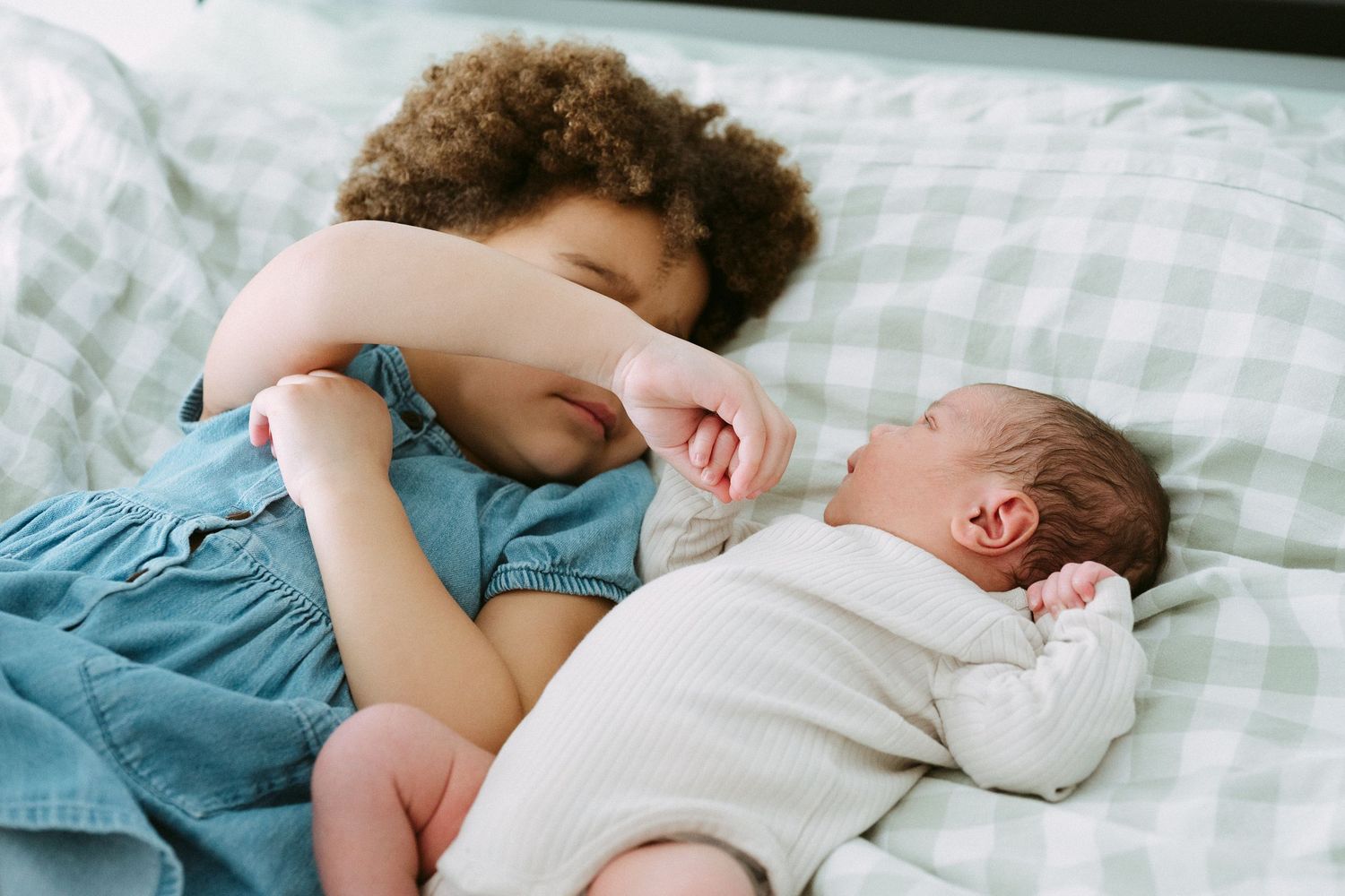 Young curly-haired child lying beside a sleeping newborn baby on a green checkered bed.