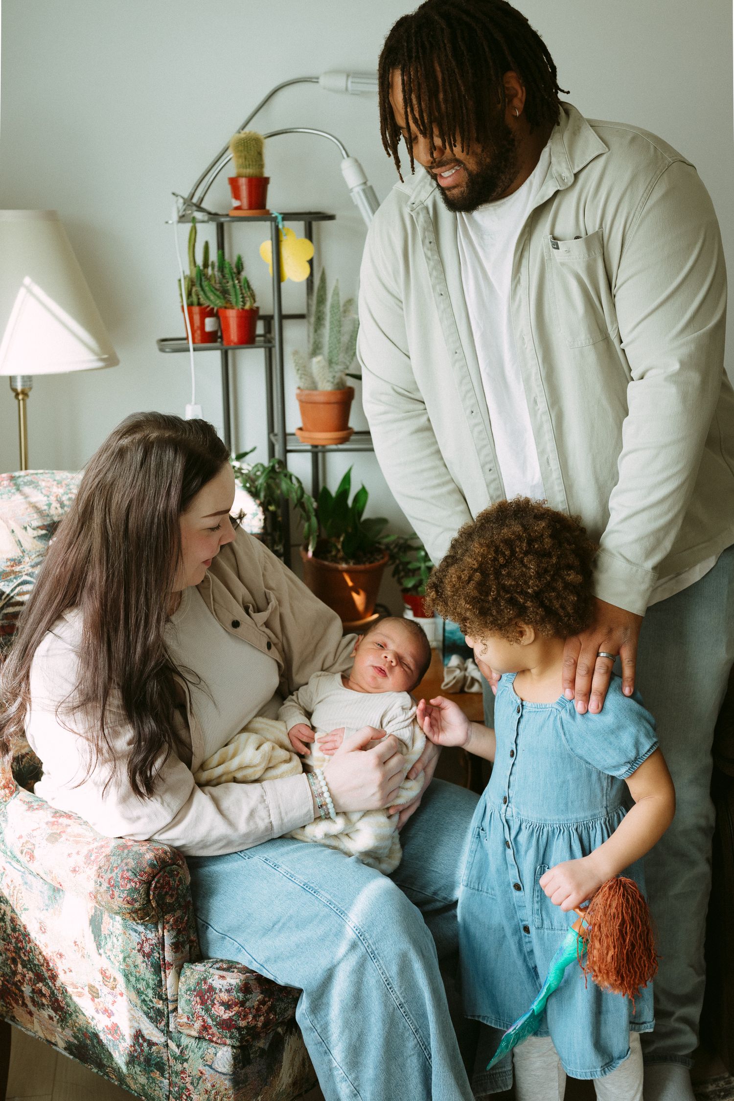 A diverse family of four admires their newborn baby indoors, surrounded by potted plants on a shelf.