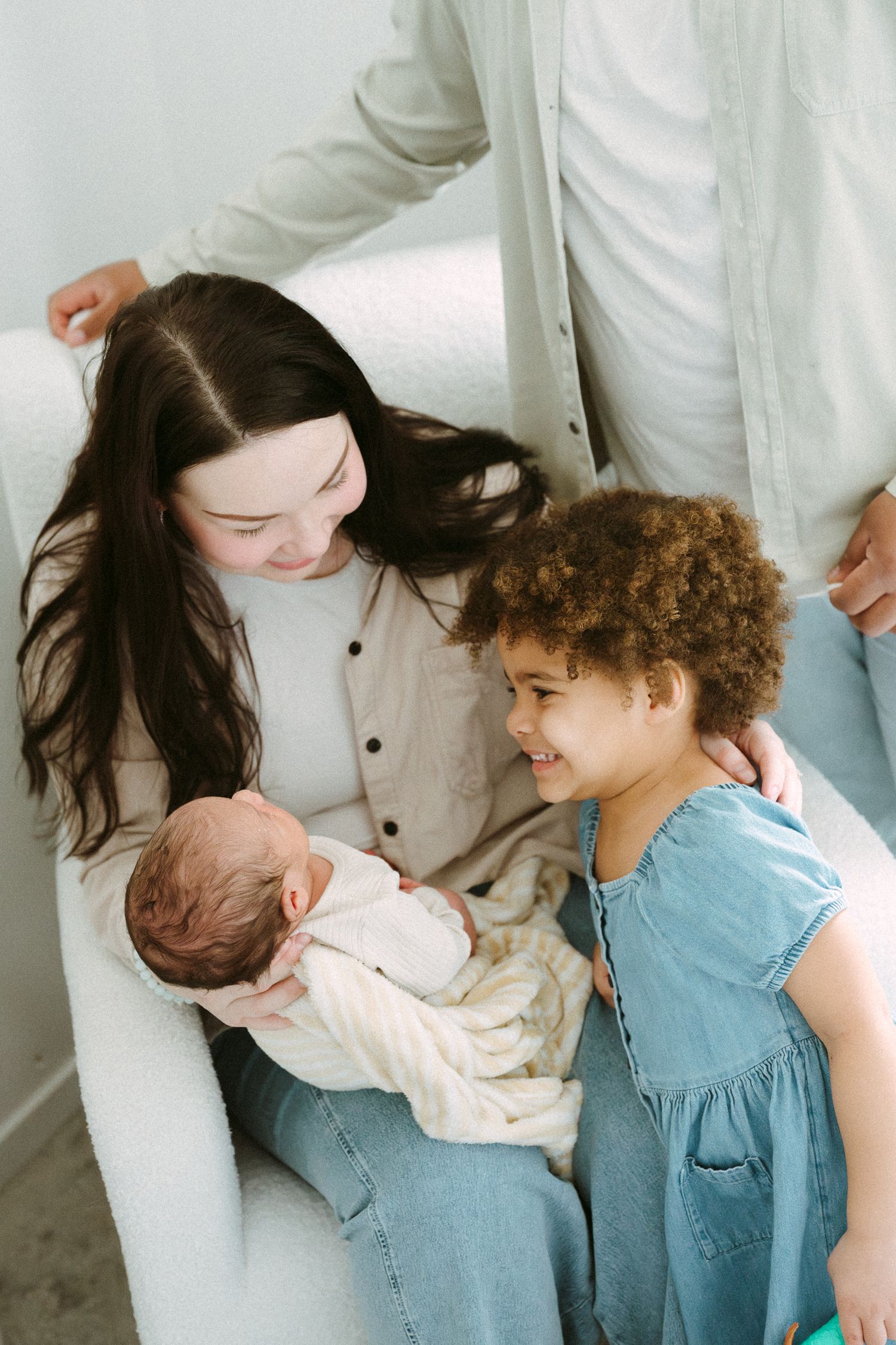 A mother with dark hair sits in a white chair holding a newborn while a curly-haired toddler girl in a blue dress smiles nearby.
