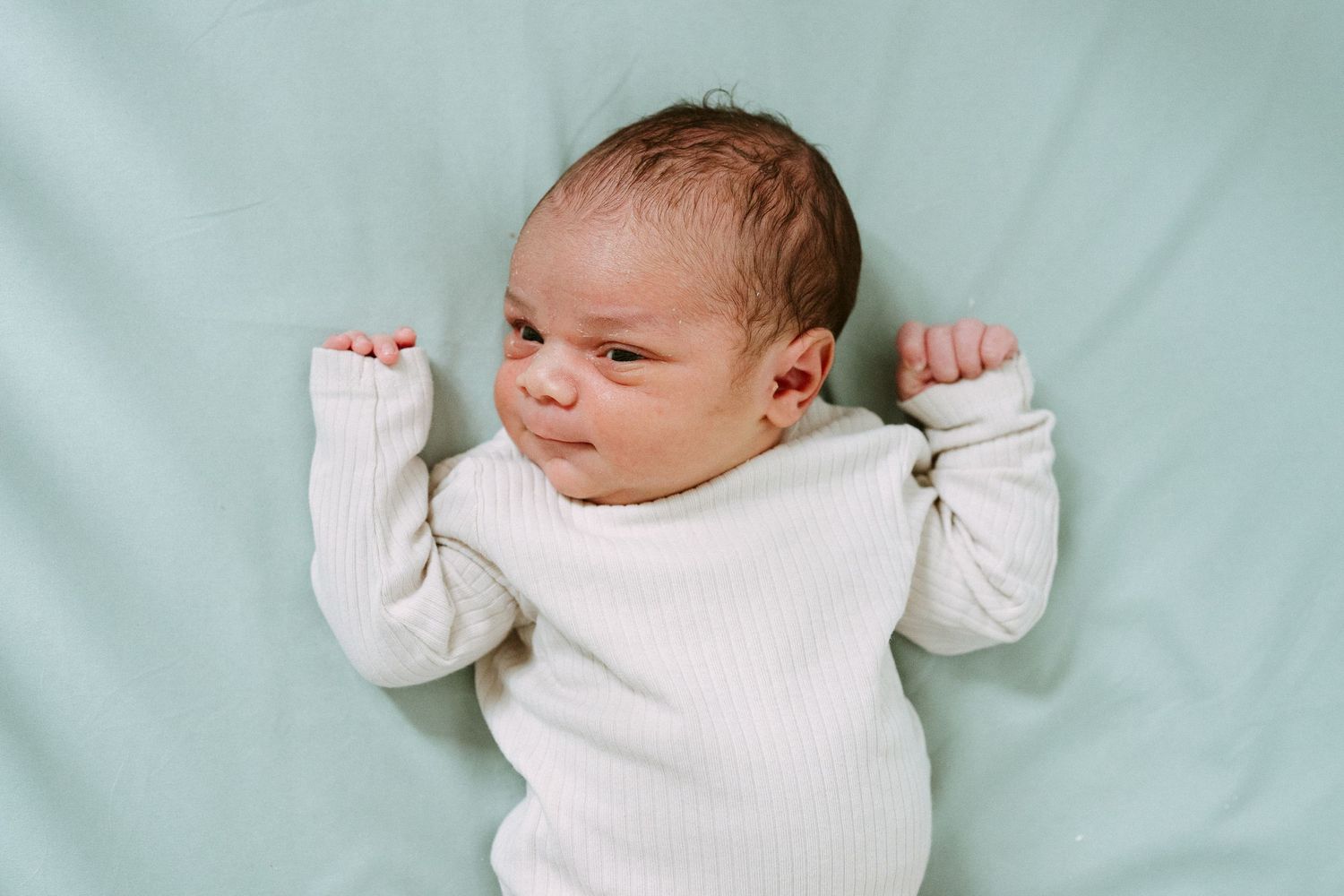 Close-up of newborn baby in white ribbed onesie lying on mint green blanket with arms raised.