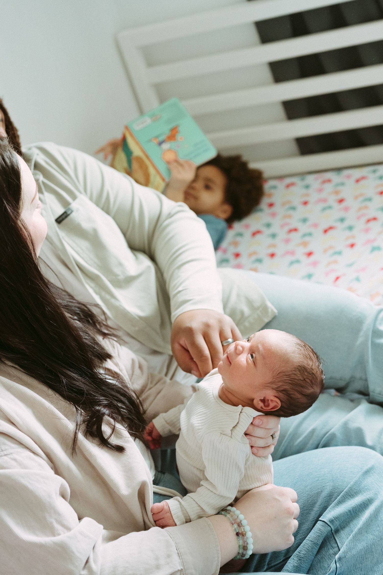 A mother tenderly holds her sleeping newborn while a child reads a book on the bed behind her.