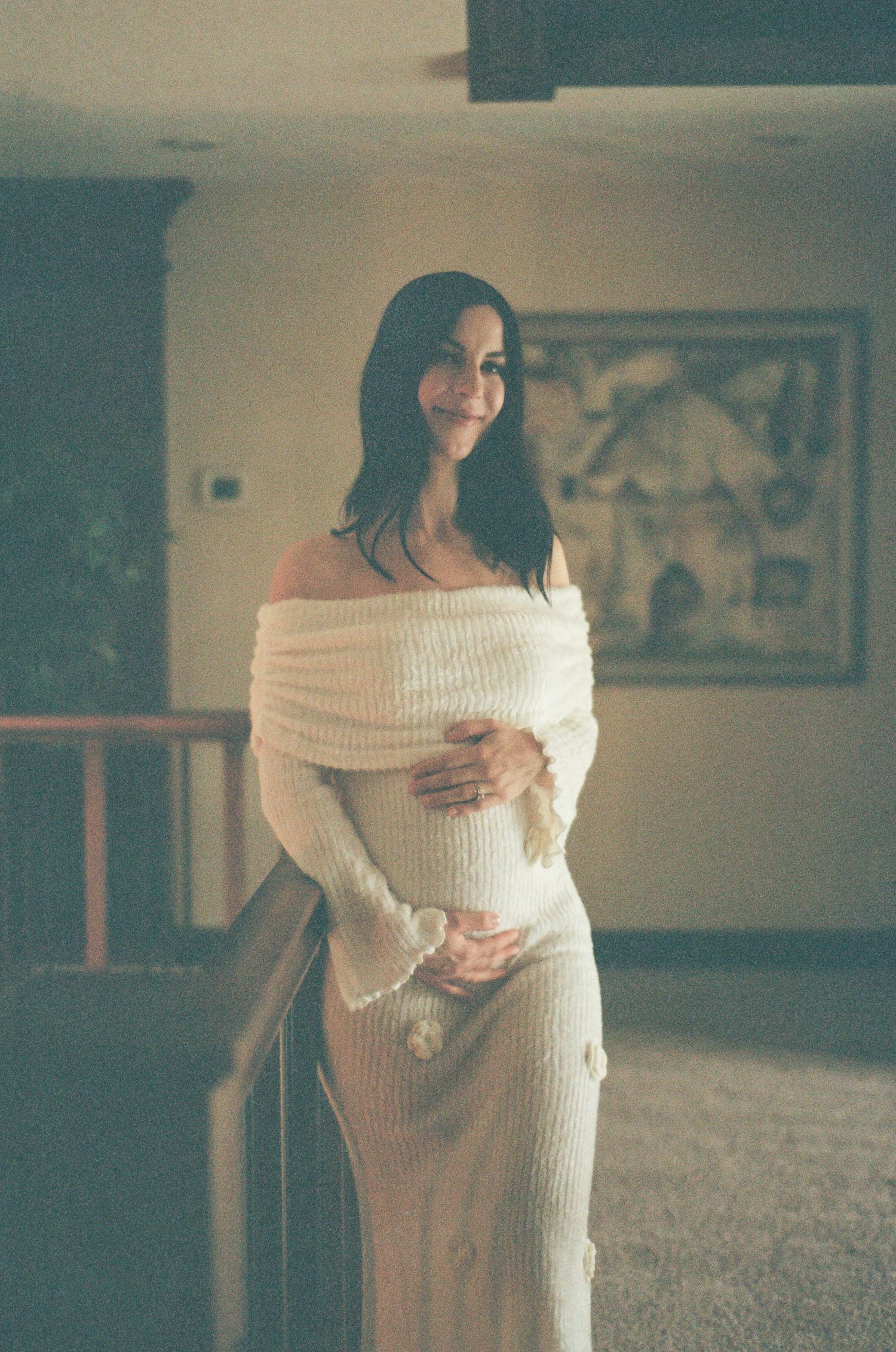 Smiling pregnant woman in white off-shoulder knit dress cradling baby bump indoors.