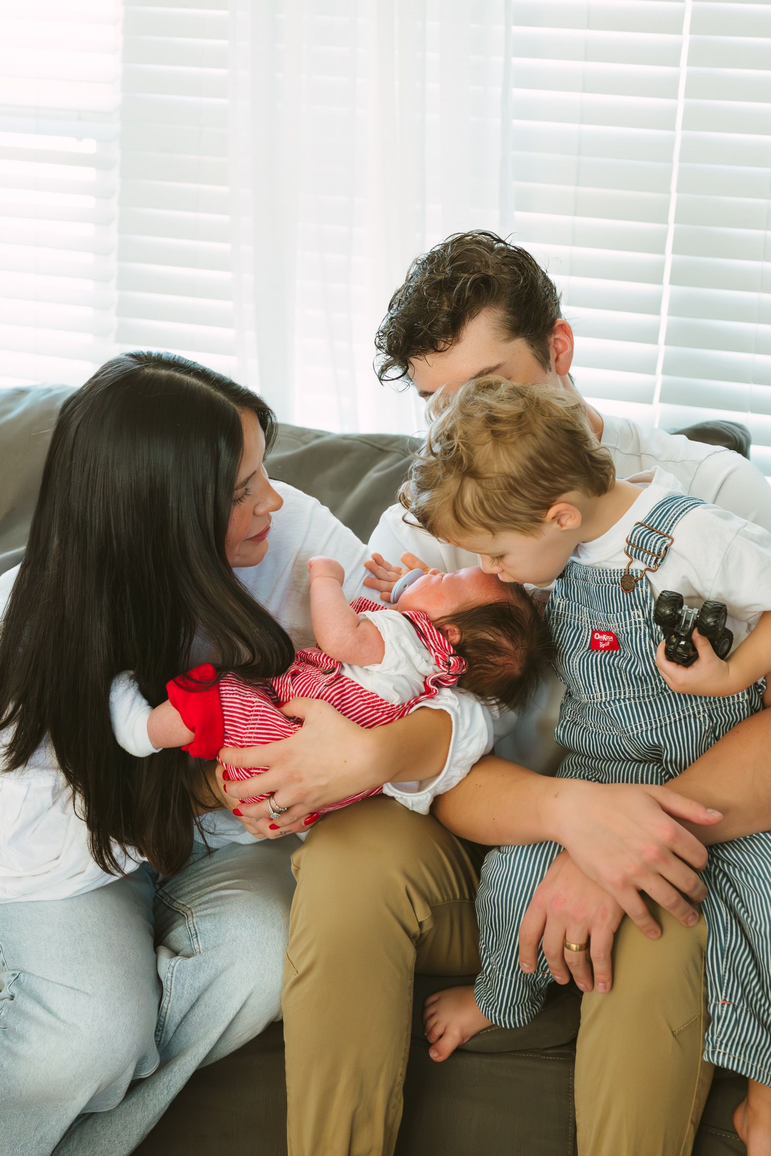 A family sits together on a gray couch, sharing a tender moment with their young children in matching outfits.