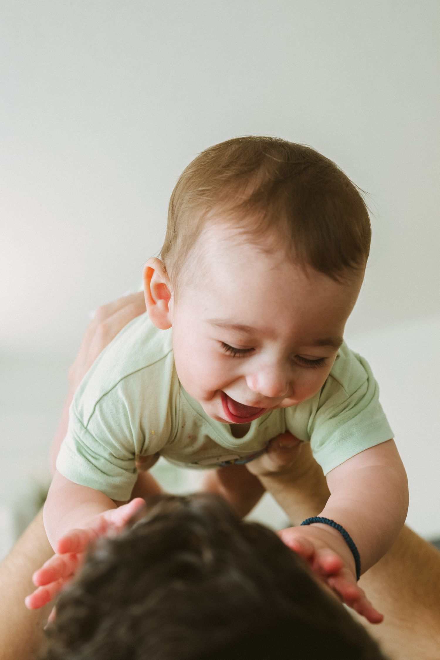 A baby in a mint green onesie smiles joyfully while being lifted up in the air.
