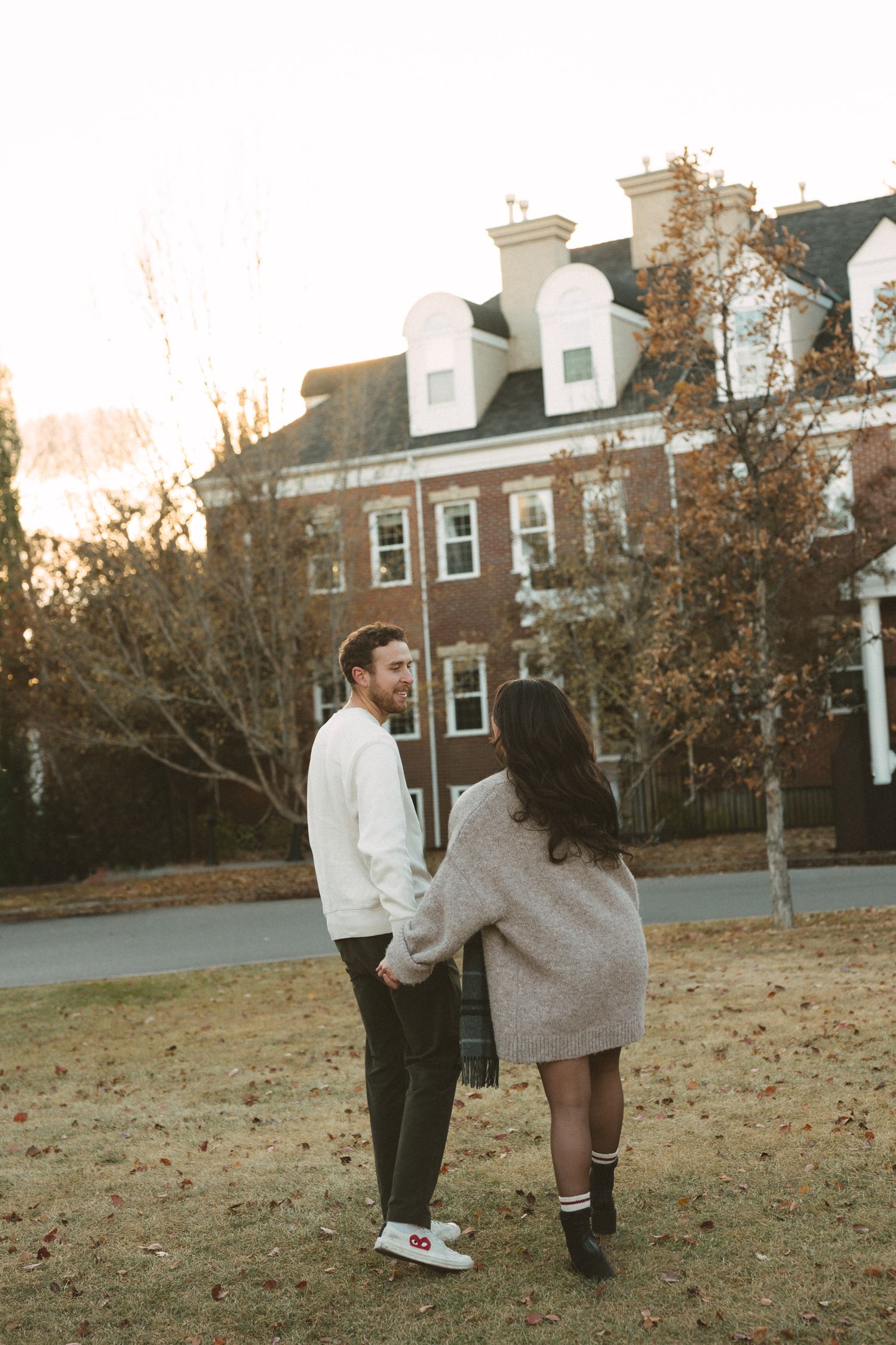 Couple playfully interacting on an autumn lawn with residential buildings visible behind them.
