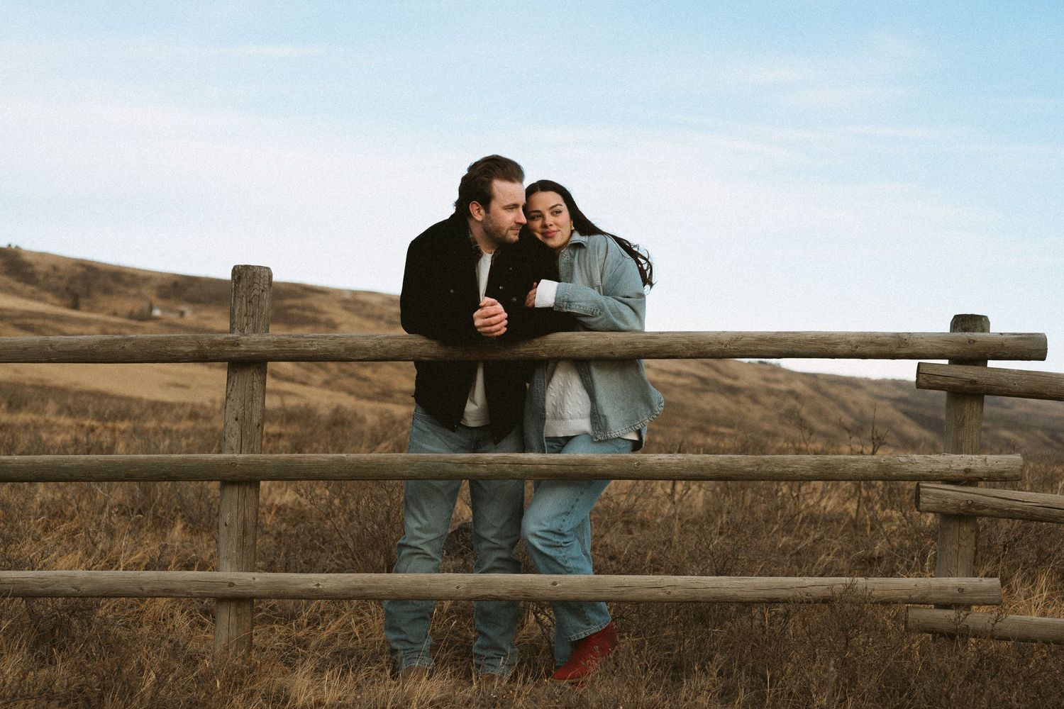 A couple embraces by a wooden fence overlooking rolling hills on a sunny day.