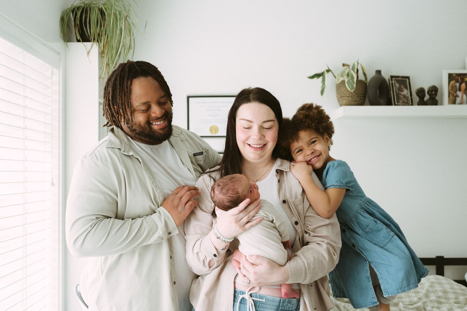 Happy interracial family smiling together, mother holding newborn baby, father and young daughter posing in bright cozy bedroom.