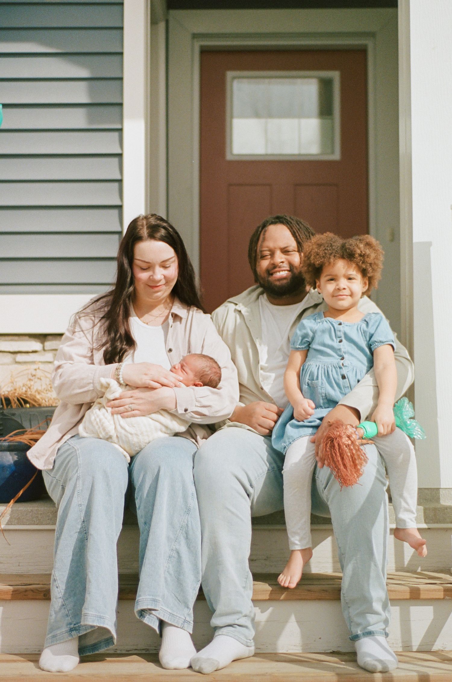 Family of four sitting on front porch steps, parents holding newborn baby and toddler girl in blue dress.