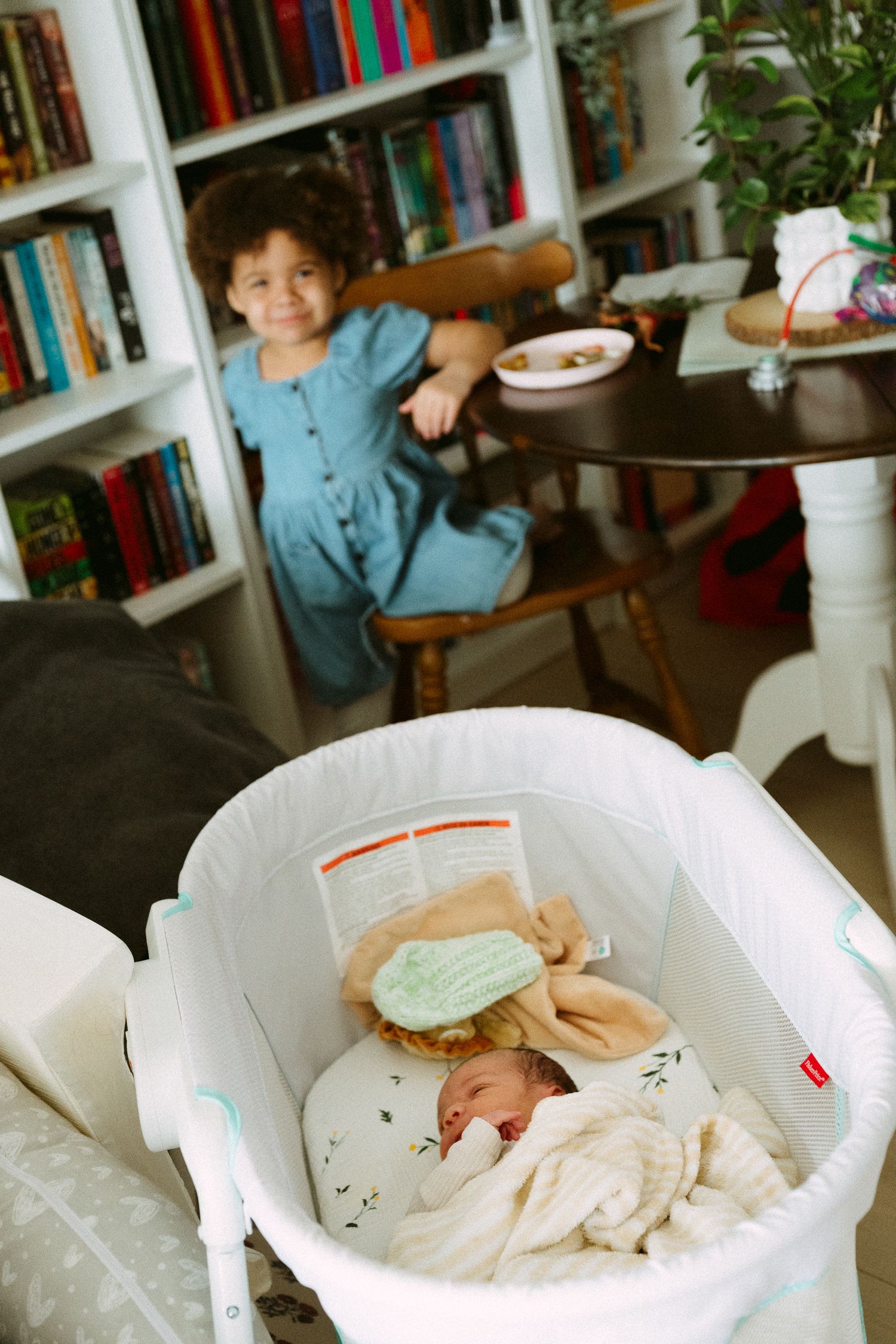 A toddler girl in a blue dress stands by a table while a newborn baby sleeps in a white bassinet nearby.