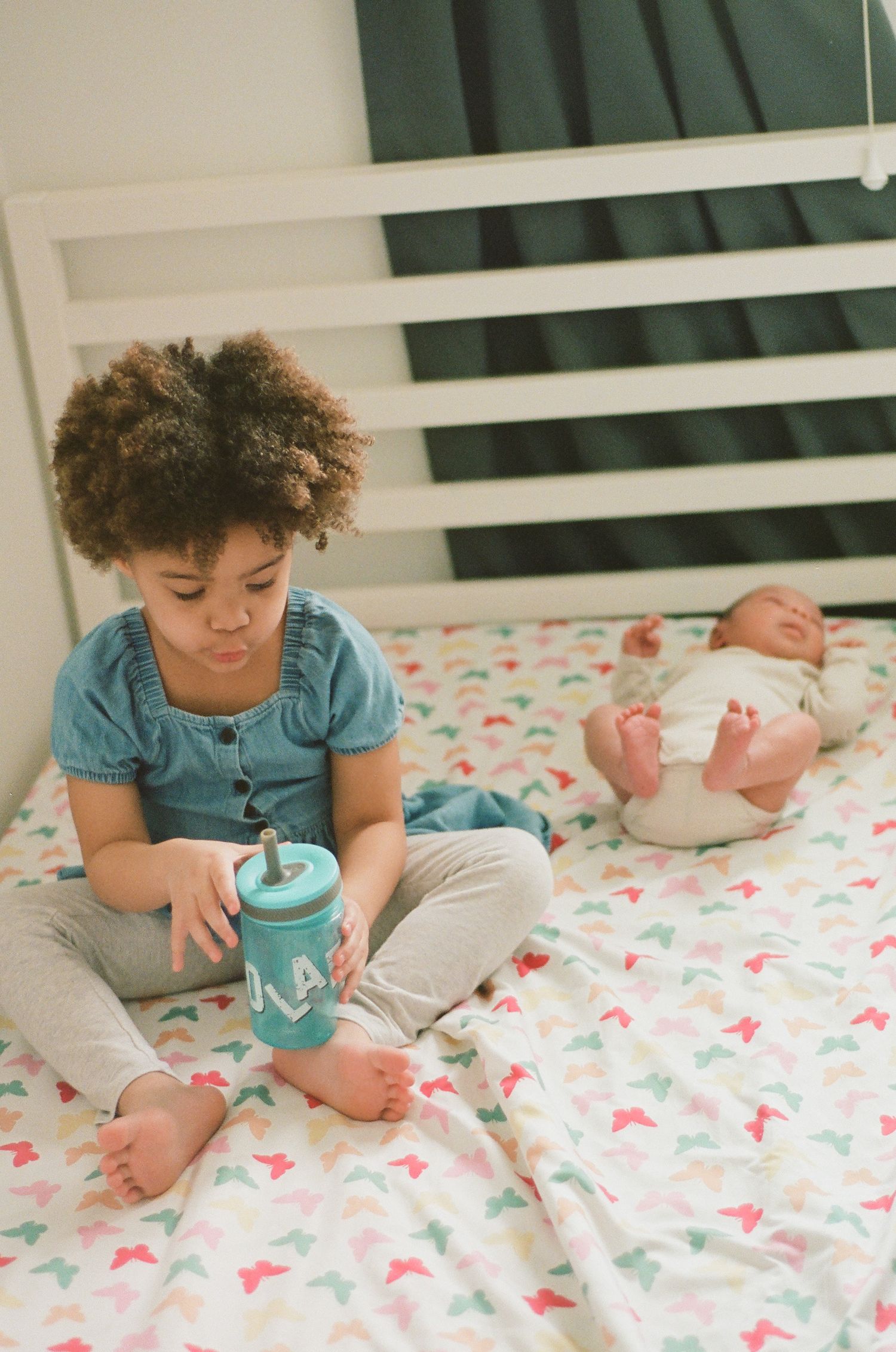 Young girl with curly hair sits on bed holding teal sippy cup beside sleeping newborn baby on colorful bedding.