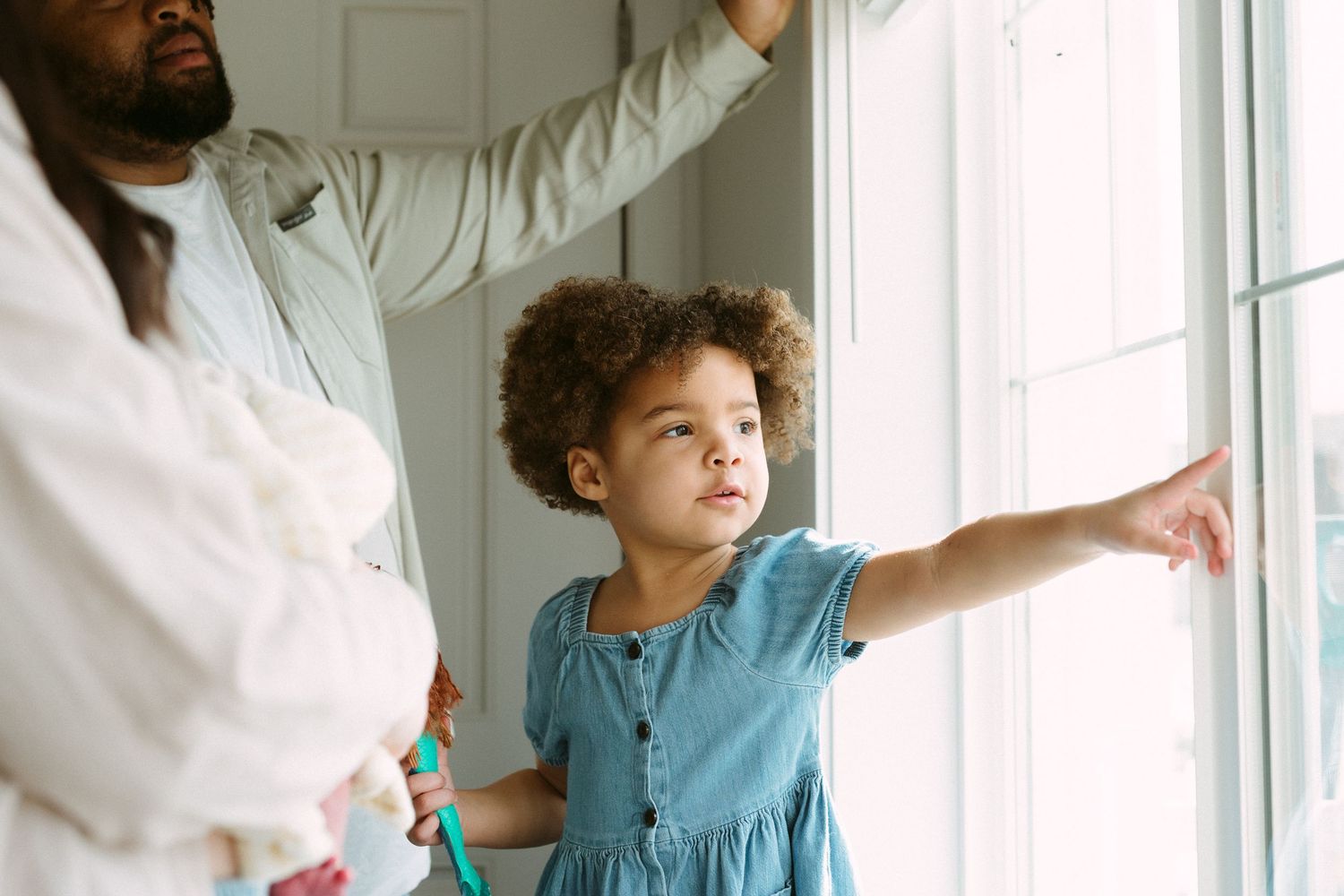 Young girl in blue dress pointing at window while adults stand nearby in bright home interior.