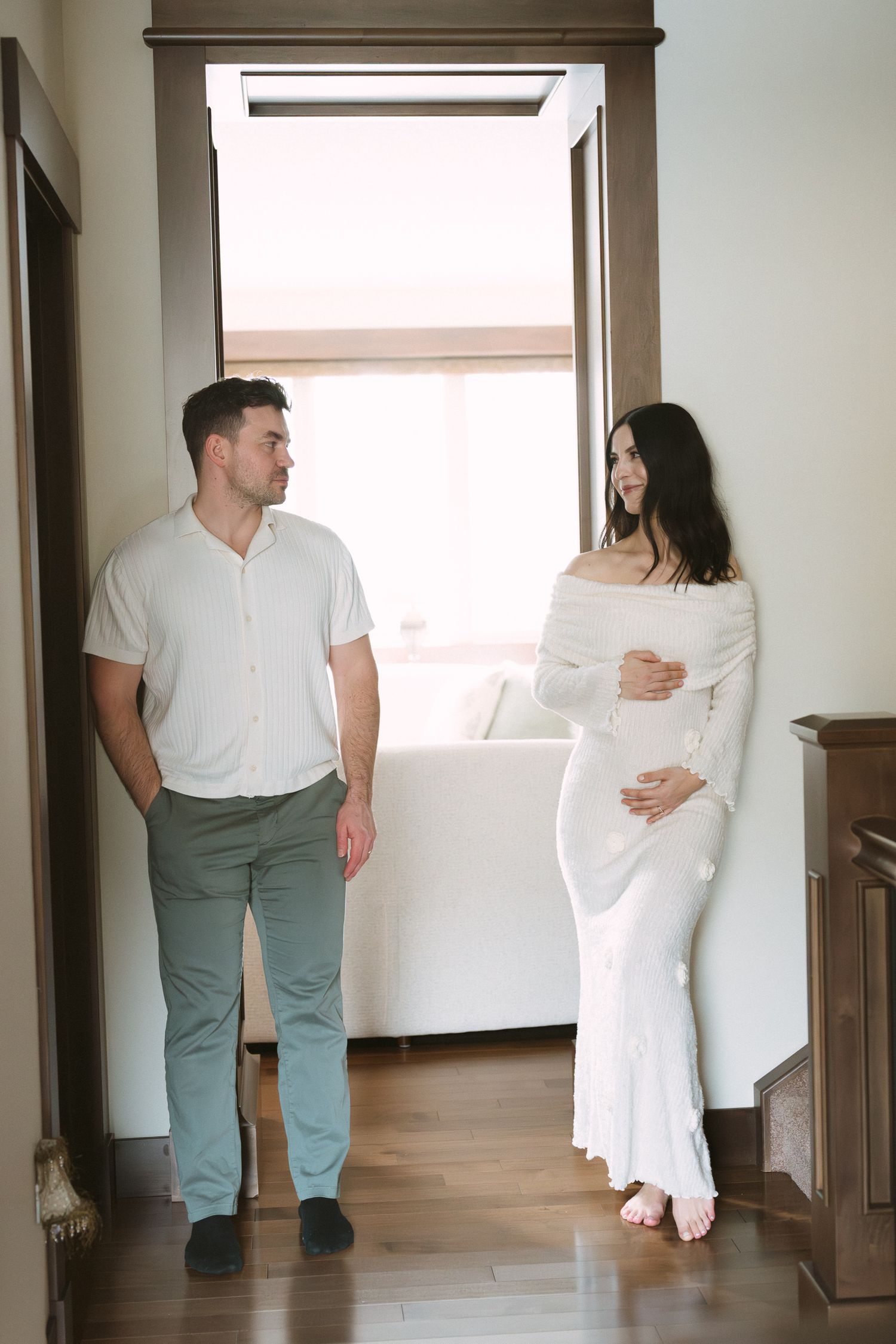 Pregnant woman in white dress holds baby bump while partner in green pants looks on indoors.