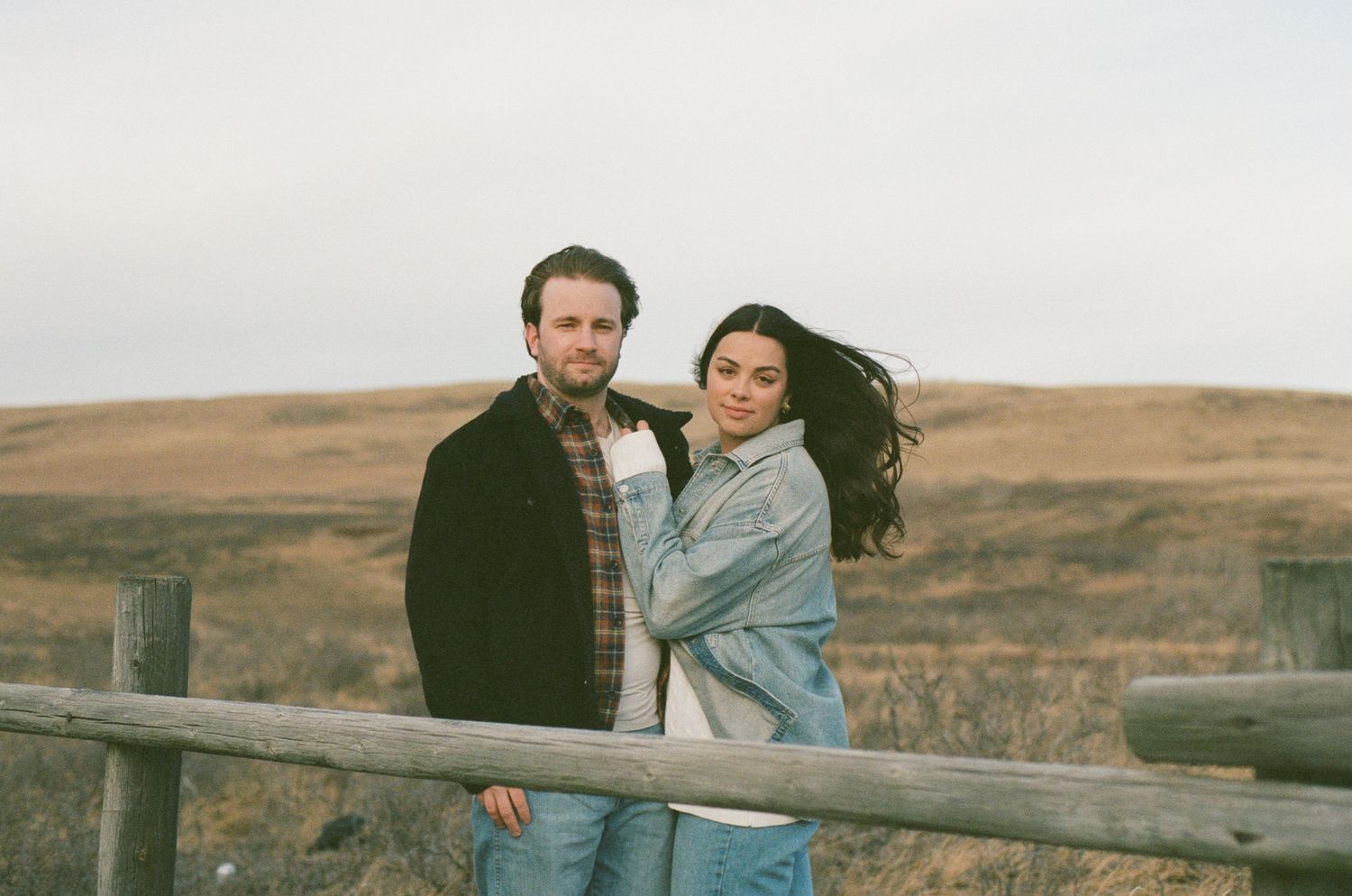 A couple embraces while standing on a wooden fence overlooking a vast autumn landscape.
