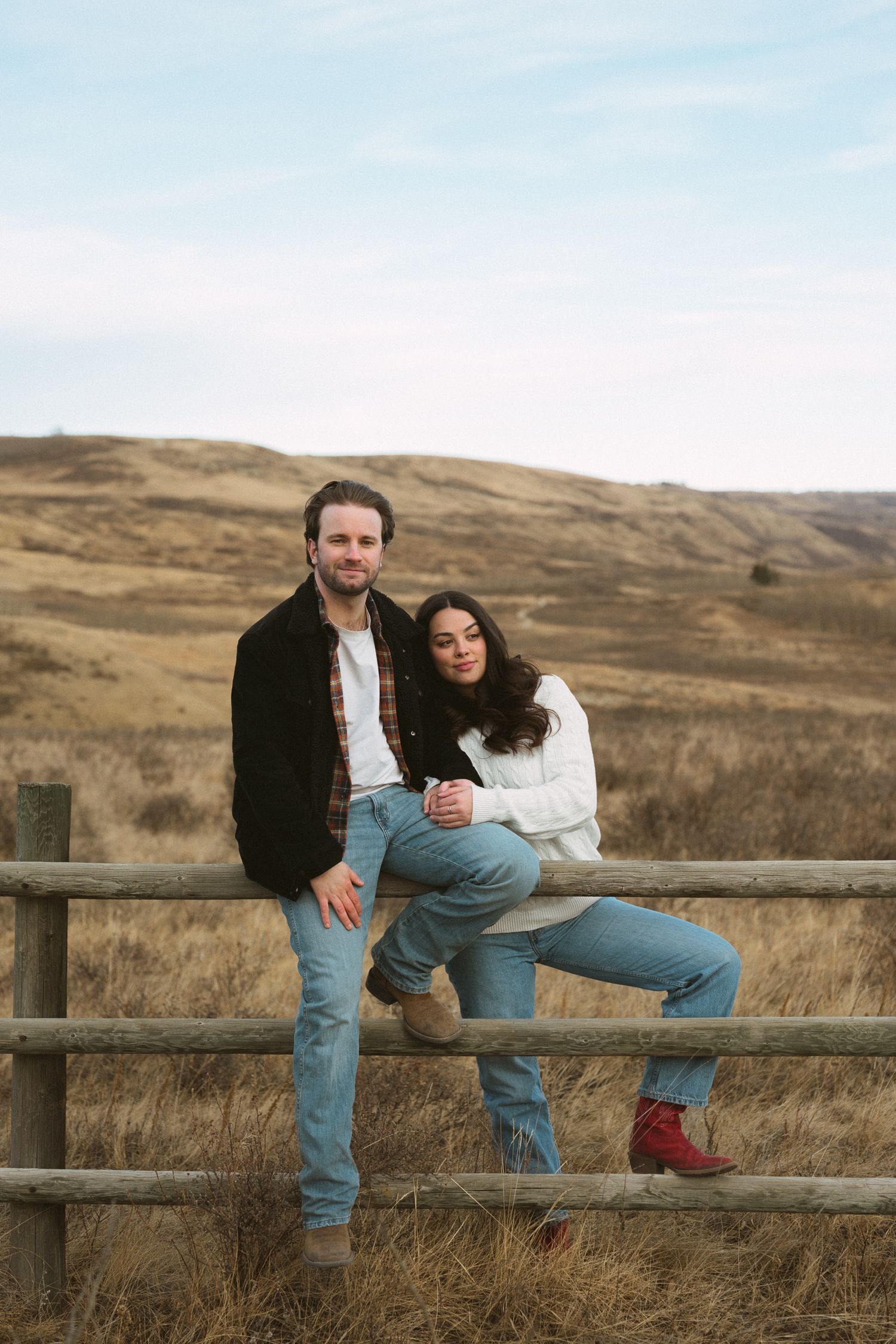A couple sits together on a wooden fence in a rustic field with rolling hills in the background.