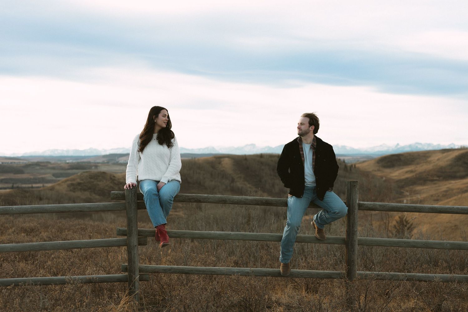 Two people share a romantic moment while sitting on a rustic wooden fence with rolling hills in the background.