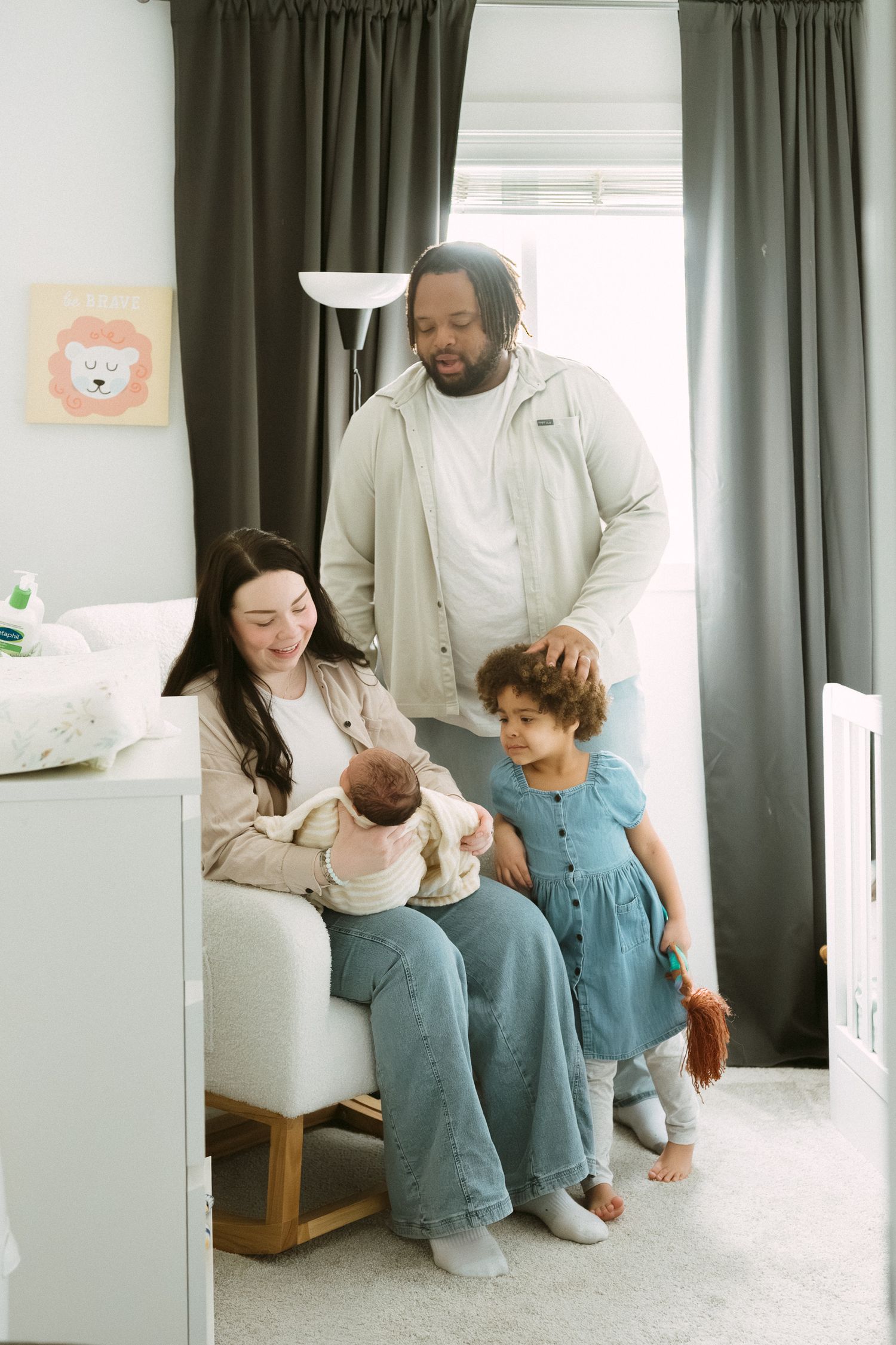 A family of four in a nursery, mother holding newborn while father and young daughter in blue dress stand beside her.
