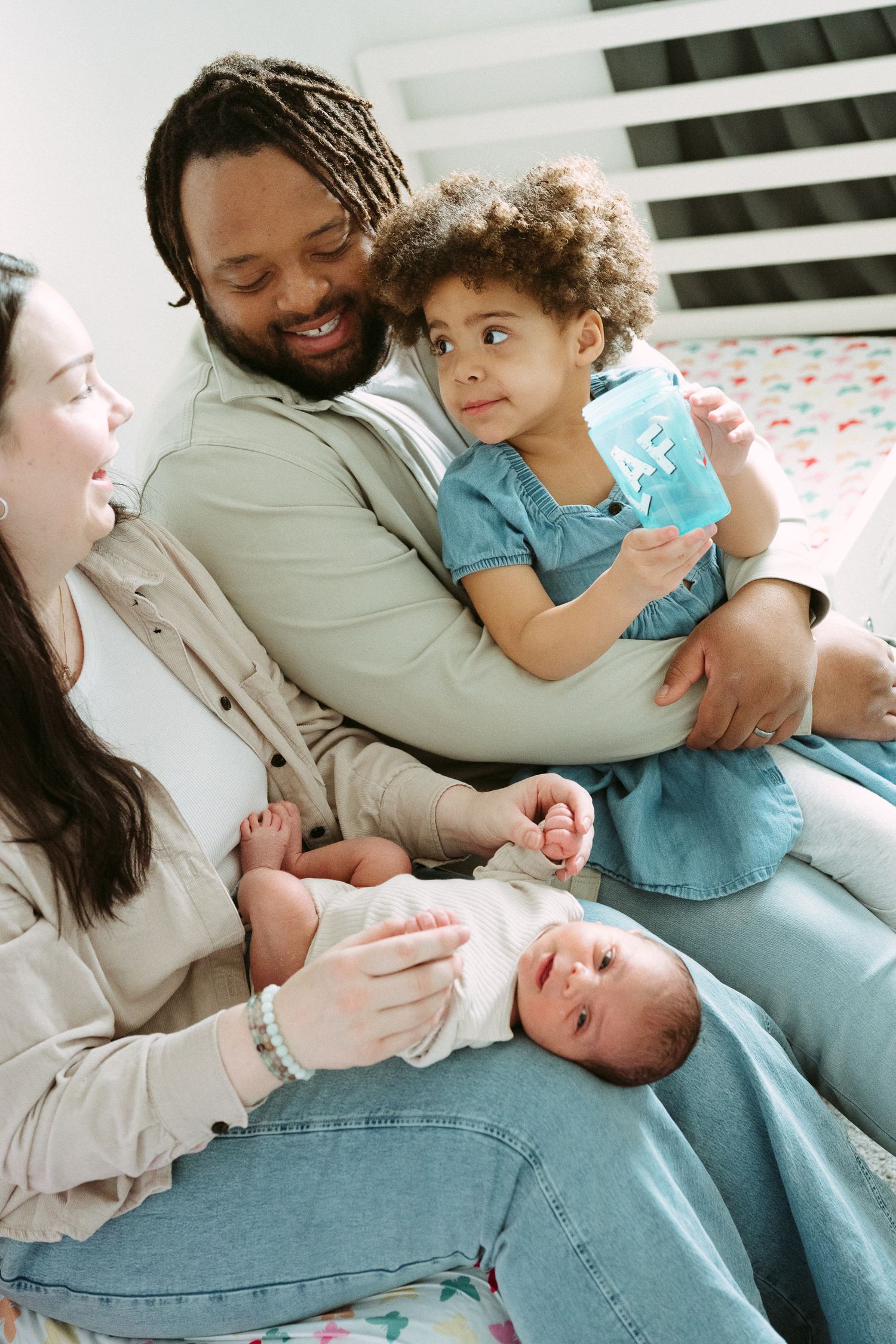 Happy multiracial family with newborn baby and toddler girl sitting together on bed at home.