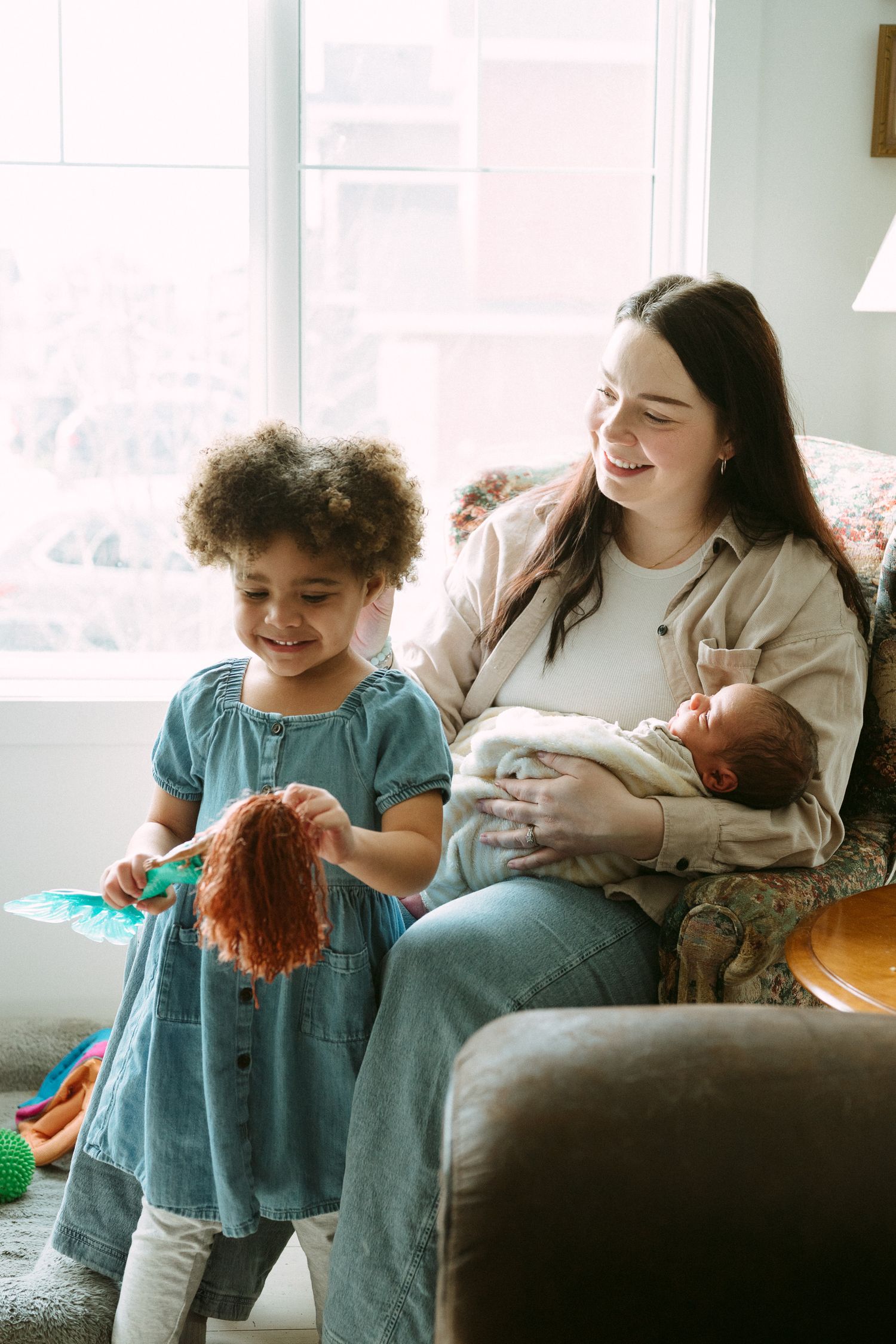 A smiling mother holds a newborn while a young girl plays with a doll in a bright, cozy living room.