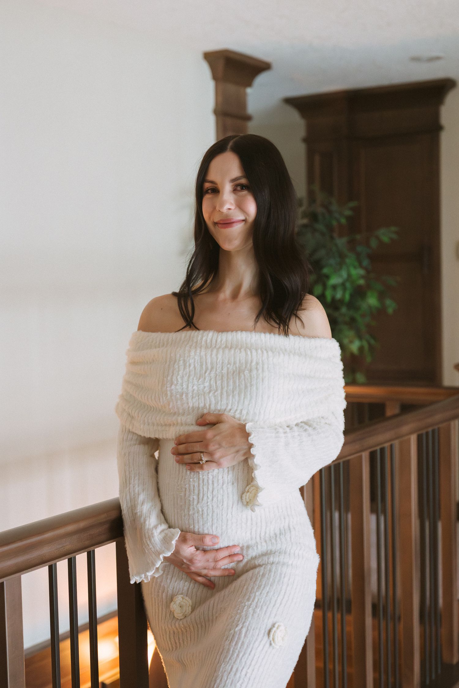 Pregnant woman in off-shoulder cream knit dress cradling baby bump, standing by wooden railing indoors.