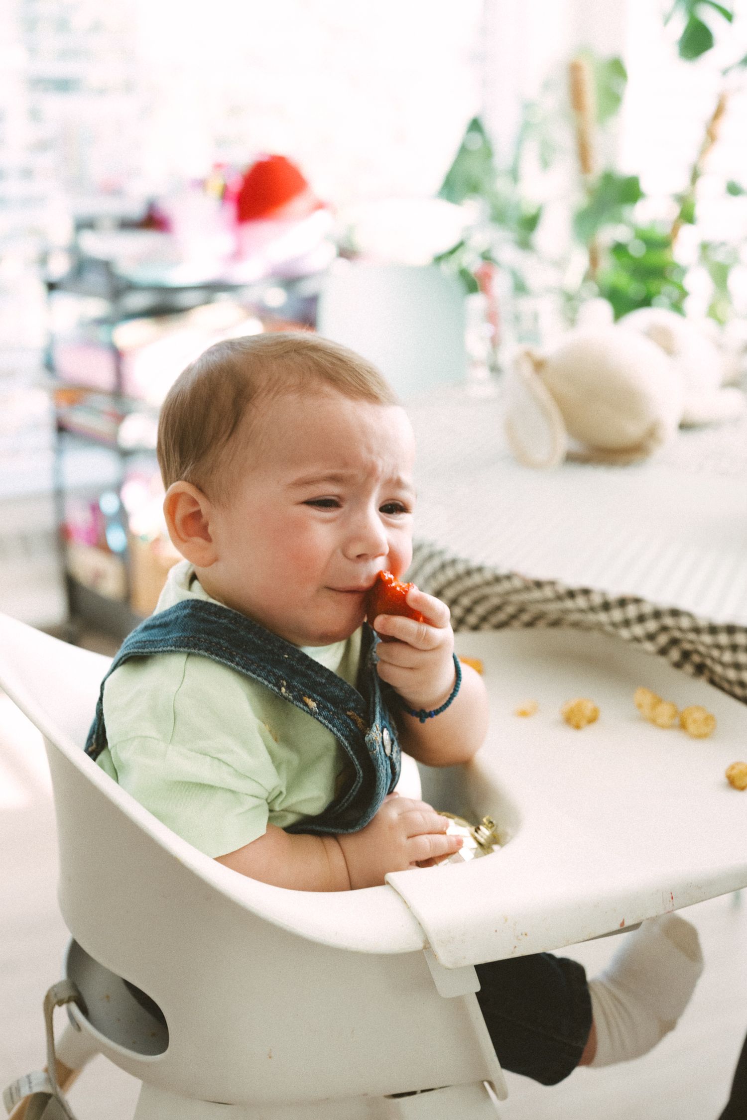 Baby in highchair eating snacks at white table.