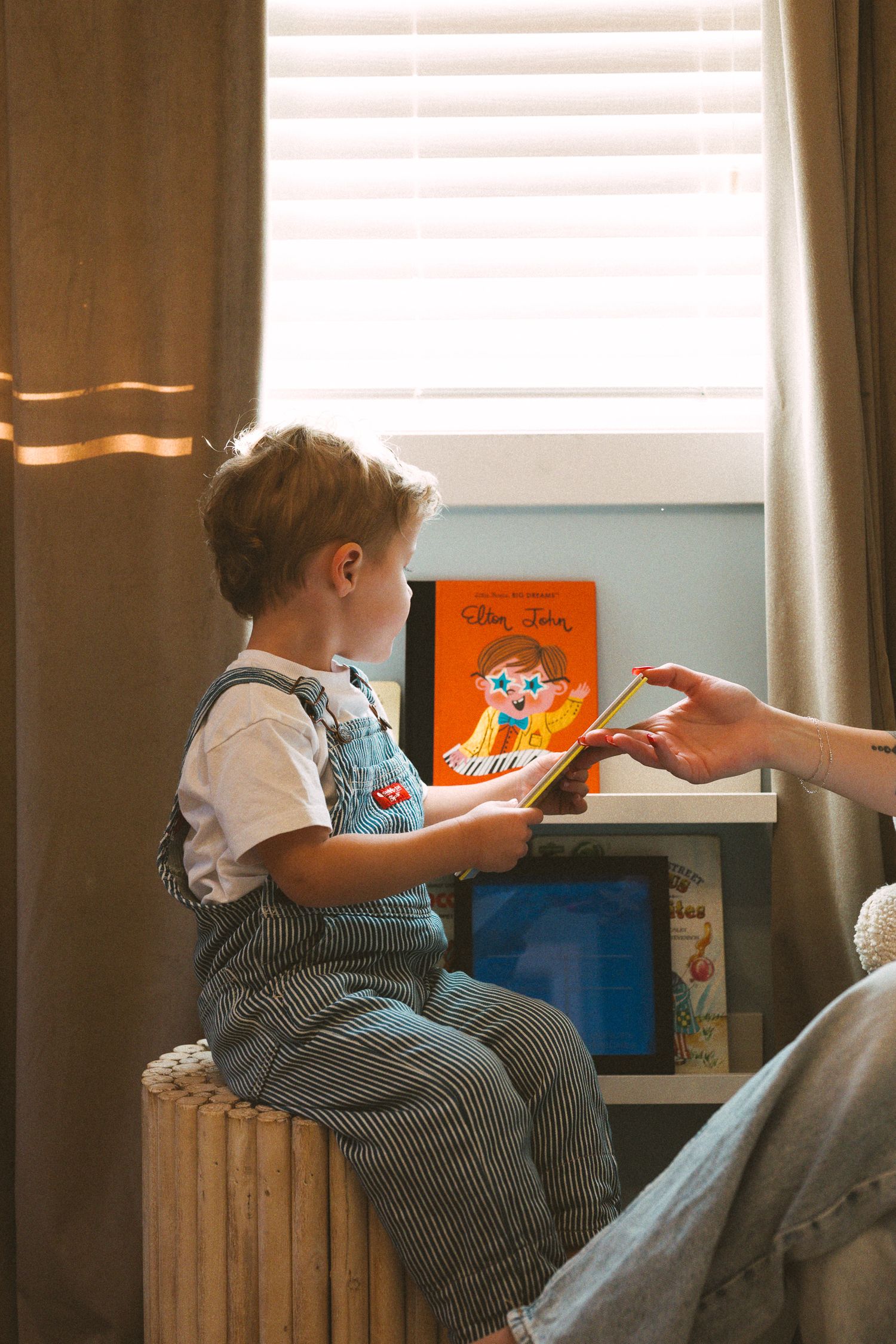 Child in striped overalls reading books while sitting on a wooden bench near a window.