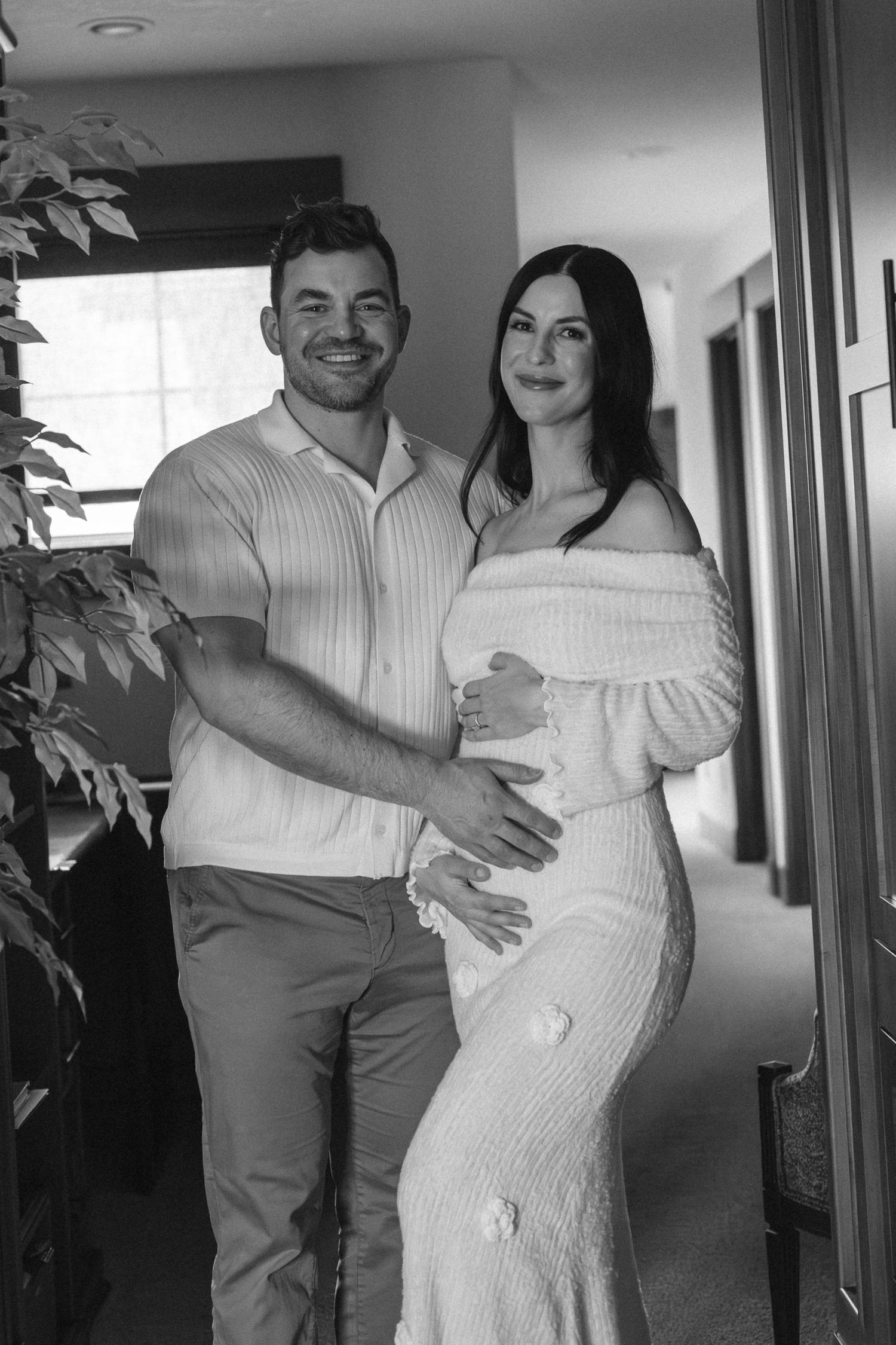 Black and white photo of a smiling couple posing together, woman in white off-shoulder maternity dress cradling baby bump.